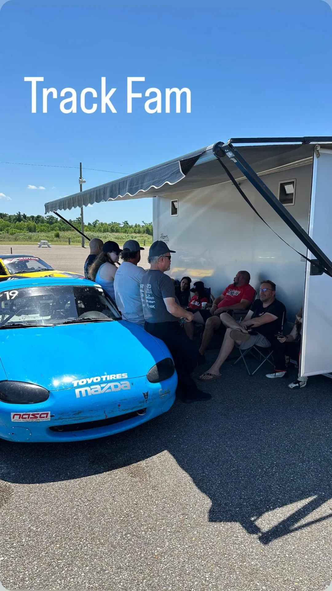 A group of people sitting under an outdoor canopy next to a racing car with racing stickers, in a parking lot on a sunny day.