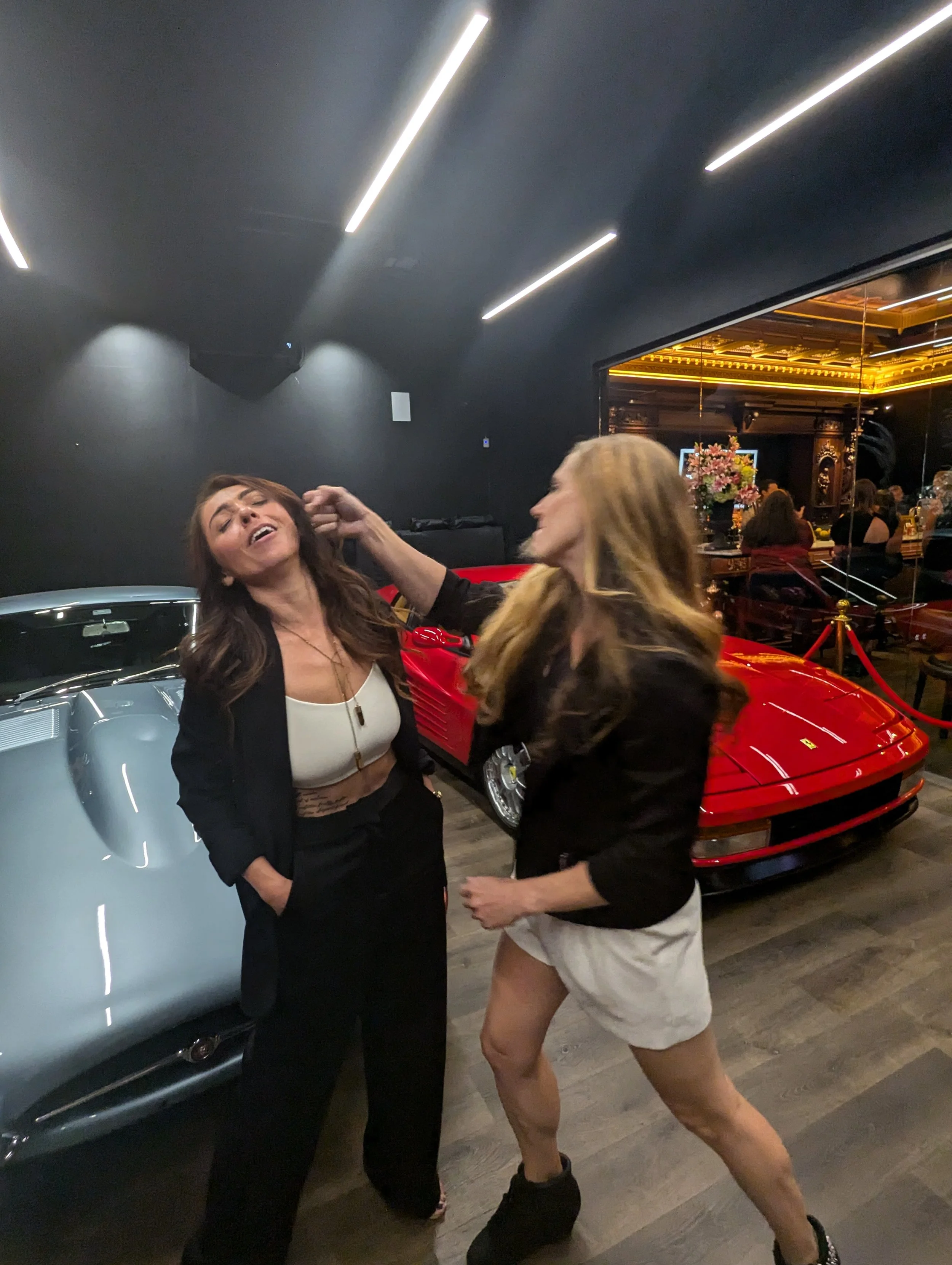 Two women are in a garage with classic cars, one woman is hitting the other on the head. There are luxury cars and a bar visible in the background.