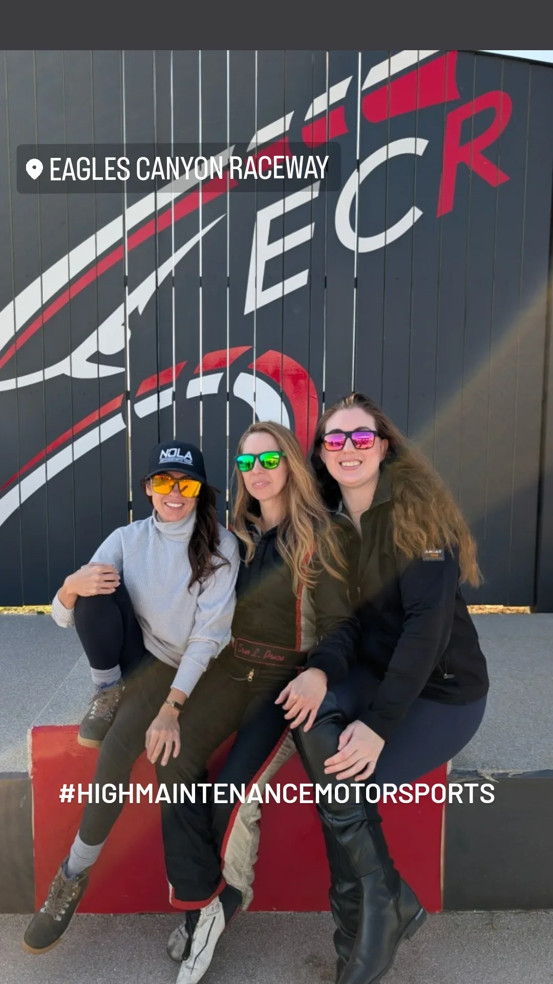 Three women wearing racing suits and sunglasses sitting together in front of a wall with a large Eagles Canyon Raceway logo. One woman is wearing a cap with 'NOLA' and all are smiling.