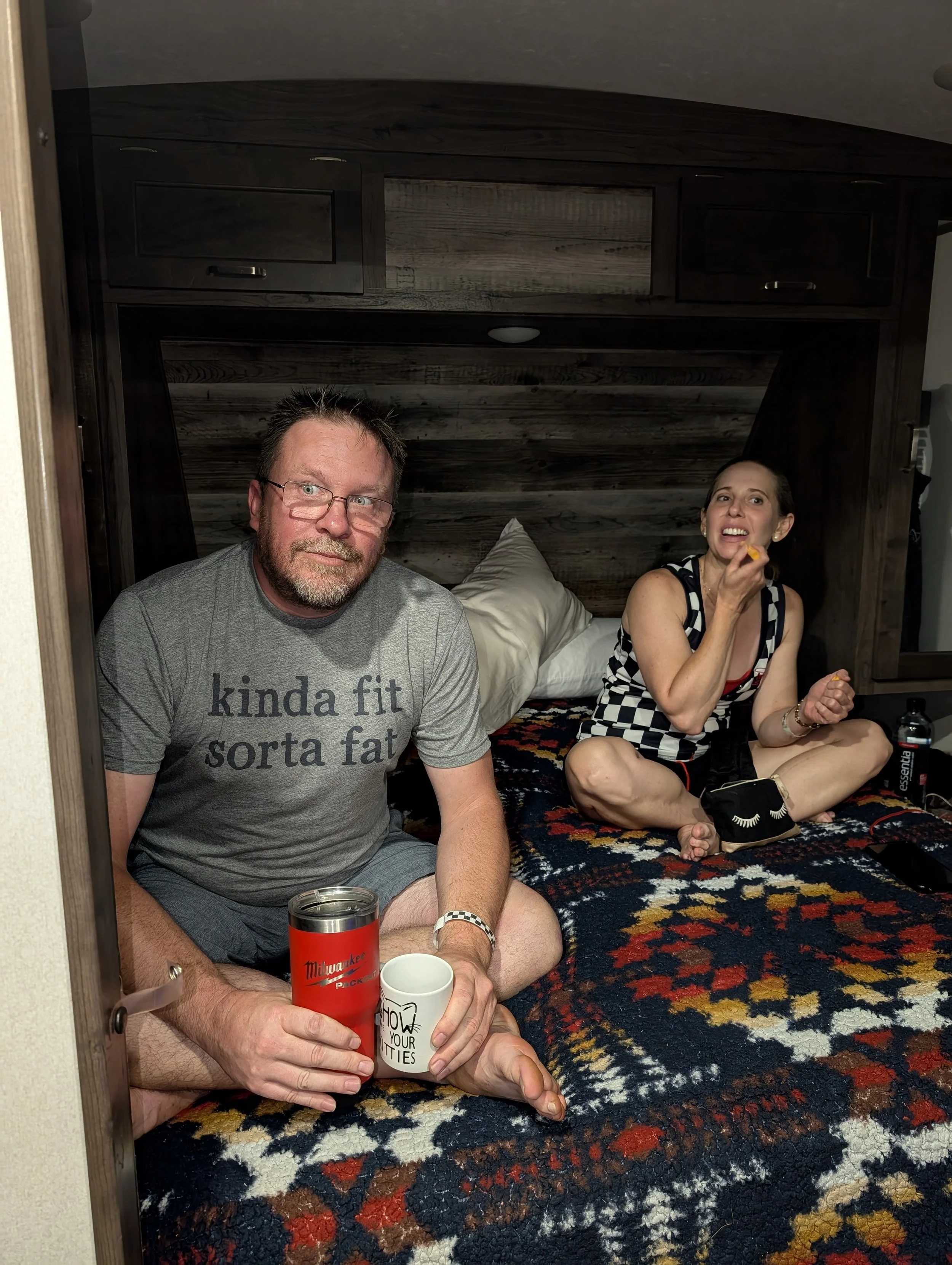 A man and a woman sitting on a bed in a cozy room. The man has glasses, a beard, and is wearing a gray T-shirt that says 'kinda fit sorta fat.' The woman is eating, wearing a black and white checkered top, and has long dark hair. The bed has a colorf