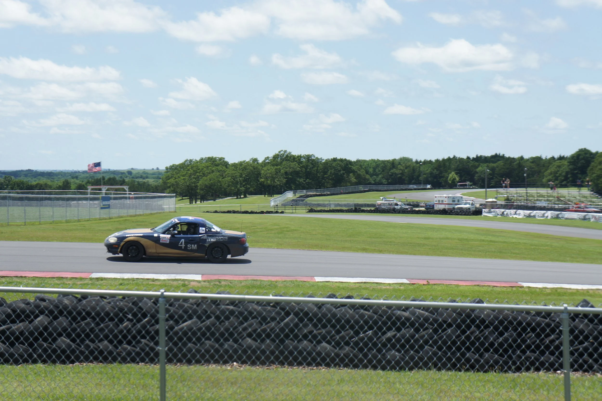 A black and gold race car on a track during a car race event on a sunny day with a blue sky and some clouds, surrounded by green grass, trees, and spectators' stands.