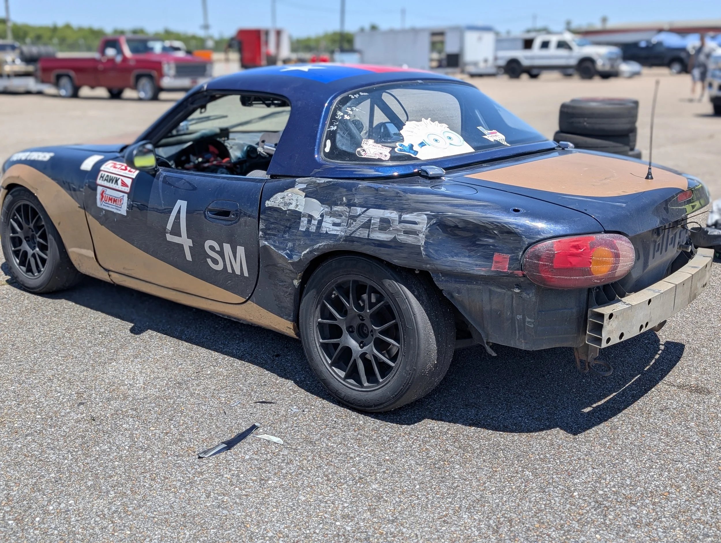A black and tan race car with the number 4 on the door, showing signs of damage, parked on a gravel lot with other vehicles in the background.