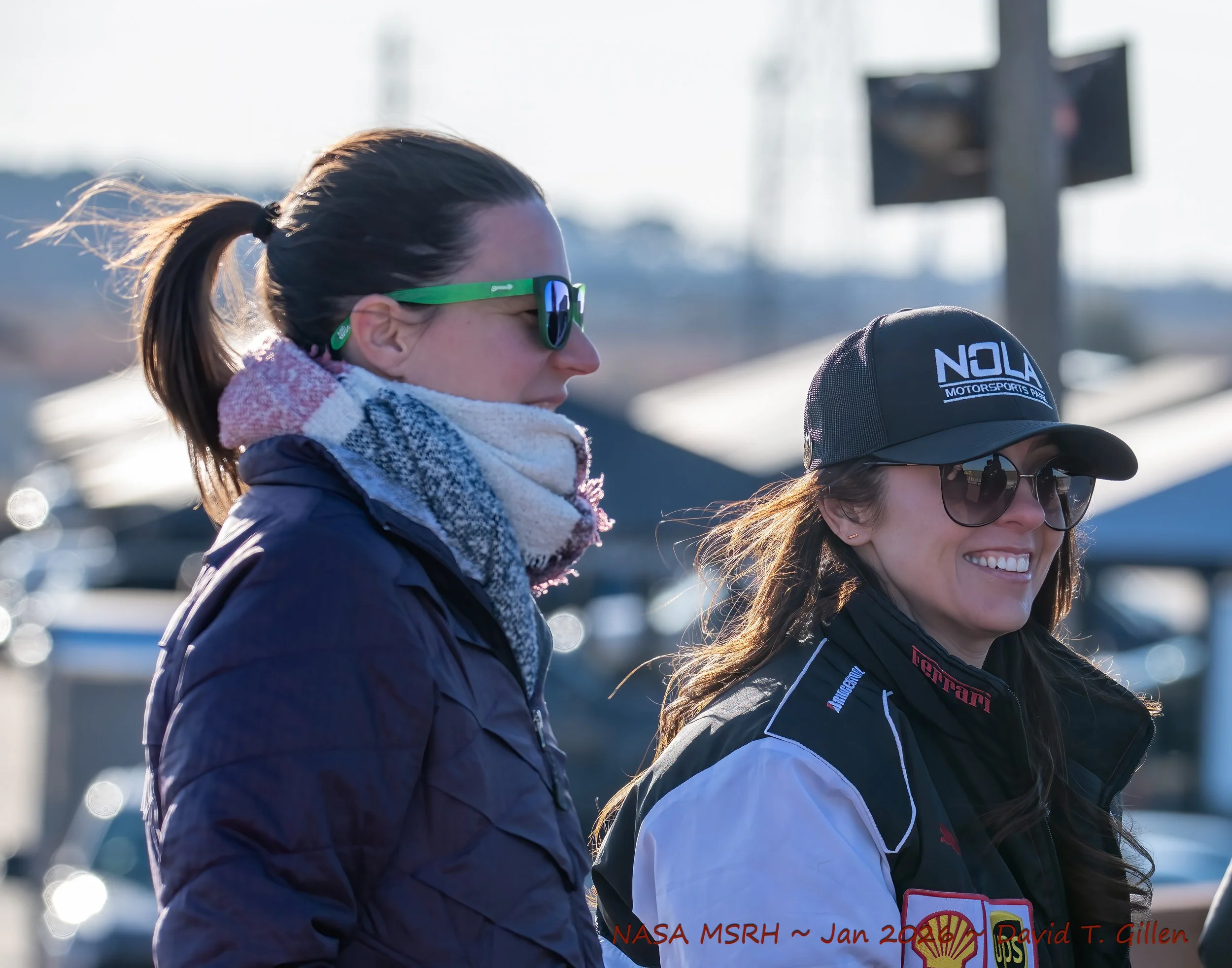 Two women standing outdoors, one in a racing jacket and cap with sponsor logos, smiling, and the other in a jacket and scarf, wearing sunglasses, with a race track in the background.
