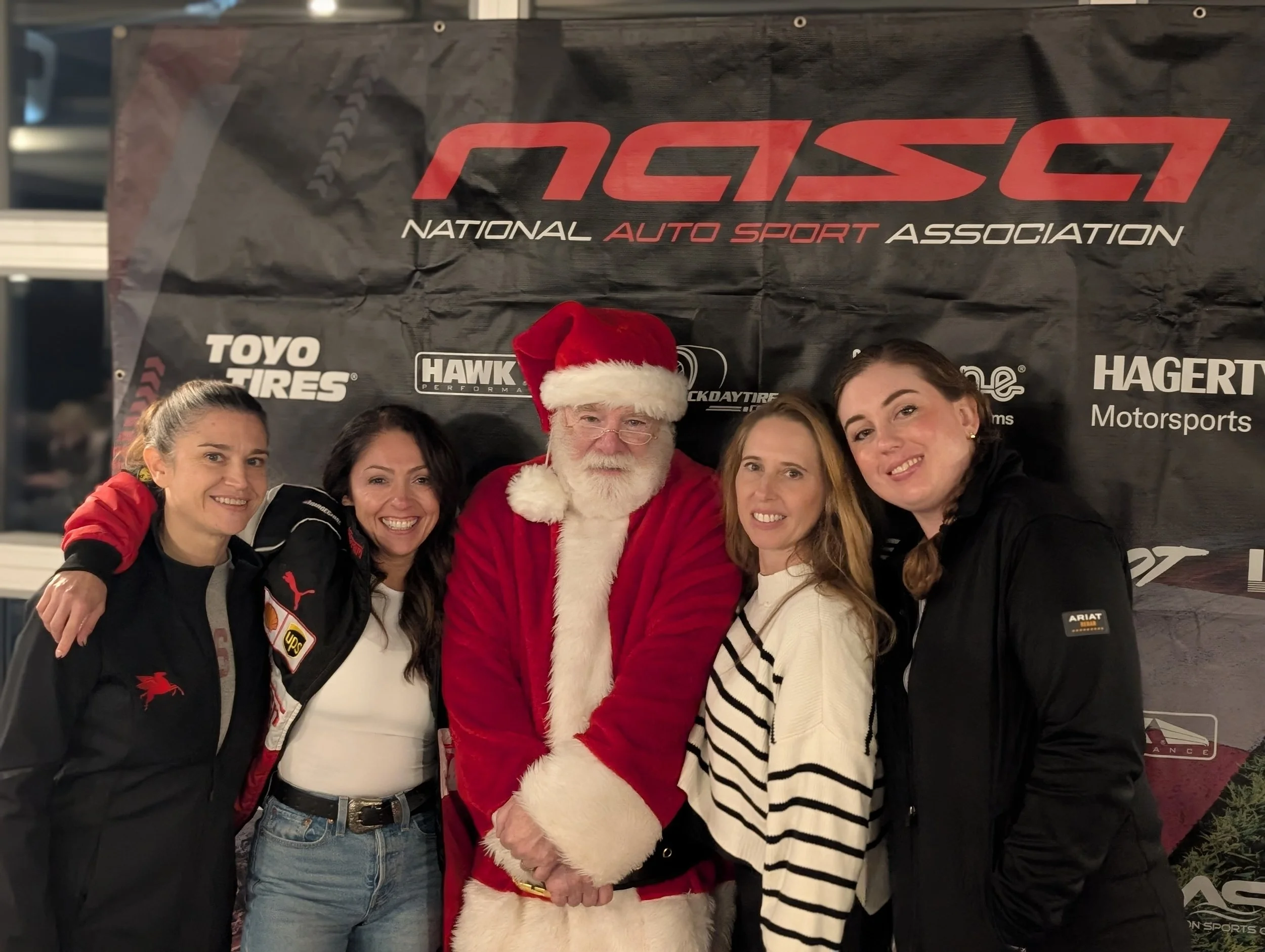 Group of four women and a man dressed as Santa Claus posing together at an auto racing event, with a black backdrop featuring logos of NOSA and various sponsors.