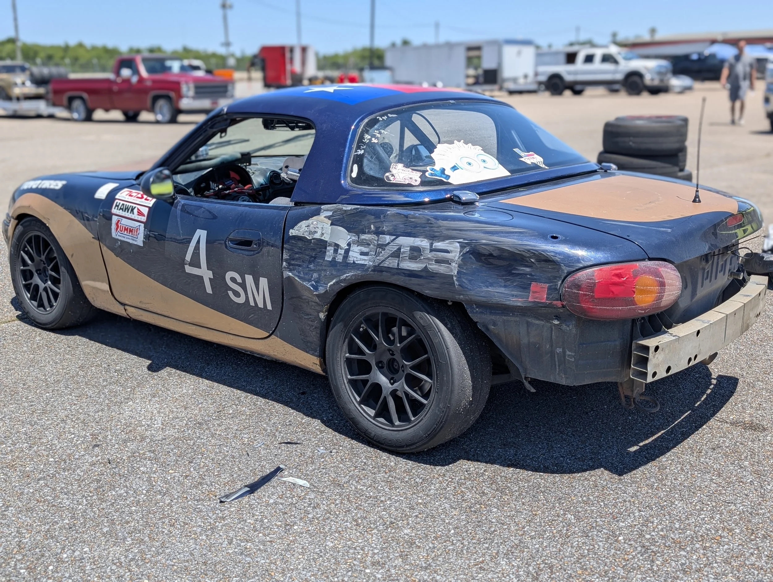 A black Mazda race car with the number 4 SM on the side, parked on a gravel surface at a race track. The car has a damaged and dirty exterior, with stickers and decals, and a SpongeBob SquarePants plush toy on the dashboard visible through the windsh