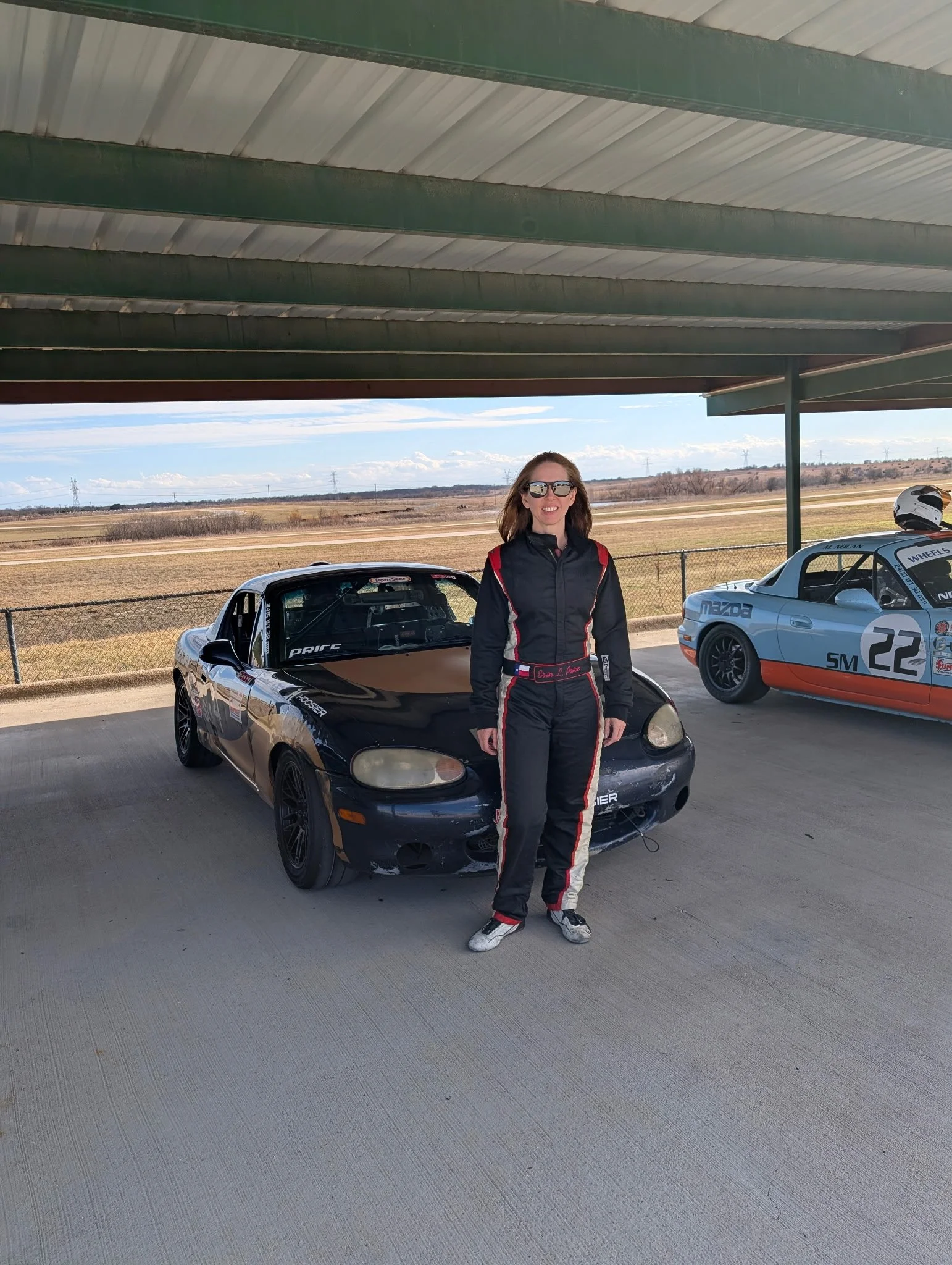 A woman in a racing suit standing next to a black race car under a shelter, with another race car in the background on a track.