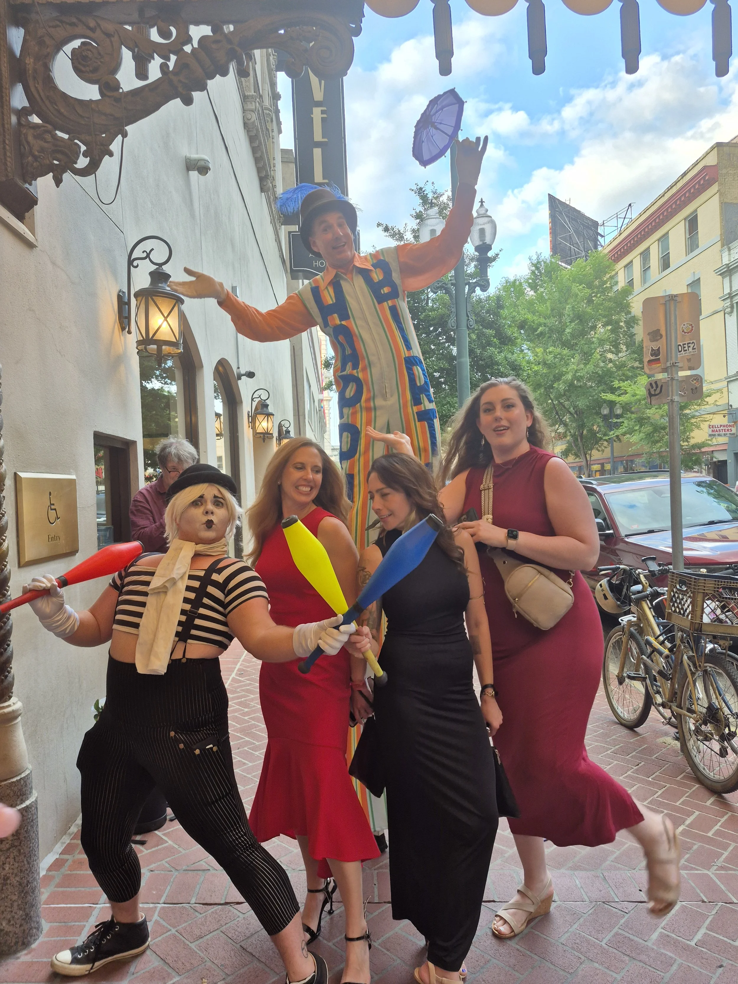 A group of four women posing with circus performers on a city sidewalk, with one performer on stilts holding an umbrella and another with a funny face holding juggling clubs, while the women strike playful poses.