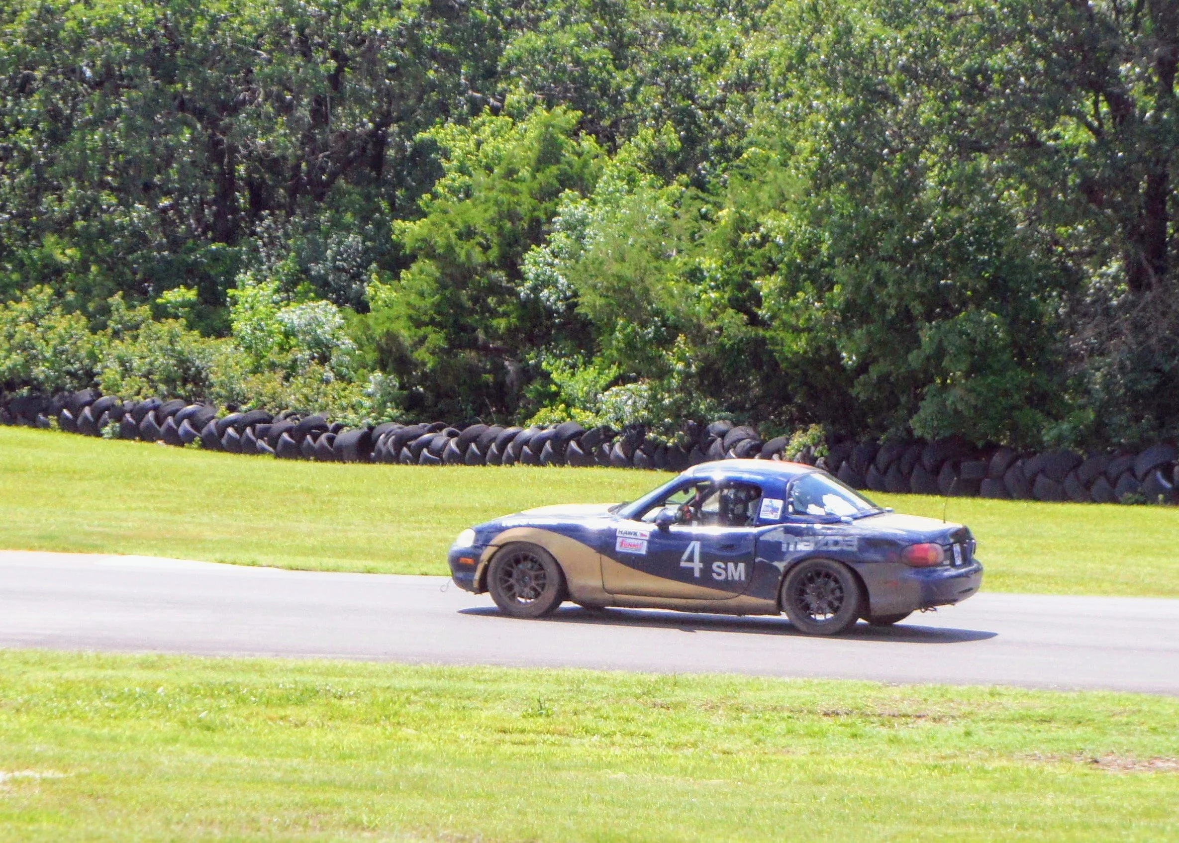 A blue racing car with the number 4 on its side driving on a racetrack with green grass and trees in the background.