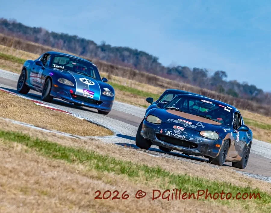Two race cars on a track, leaning into a turn, with grass and trees in the background.