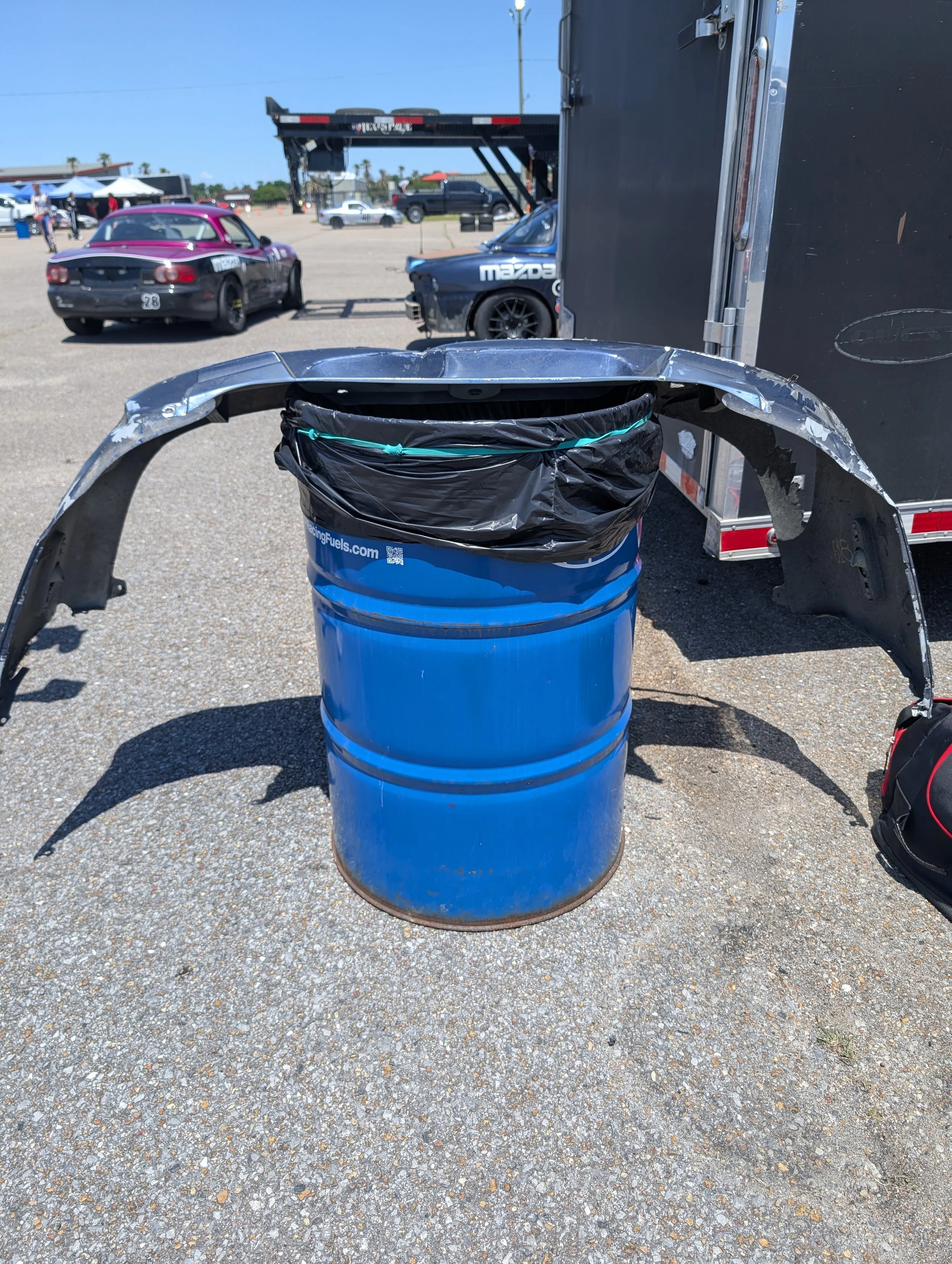 A blue barrel with a black trash bag on top, surrounded by car parts including a windshield and fenders, in an outdoor parking lot with various vehicles and tents in the background under a clear blue sky.