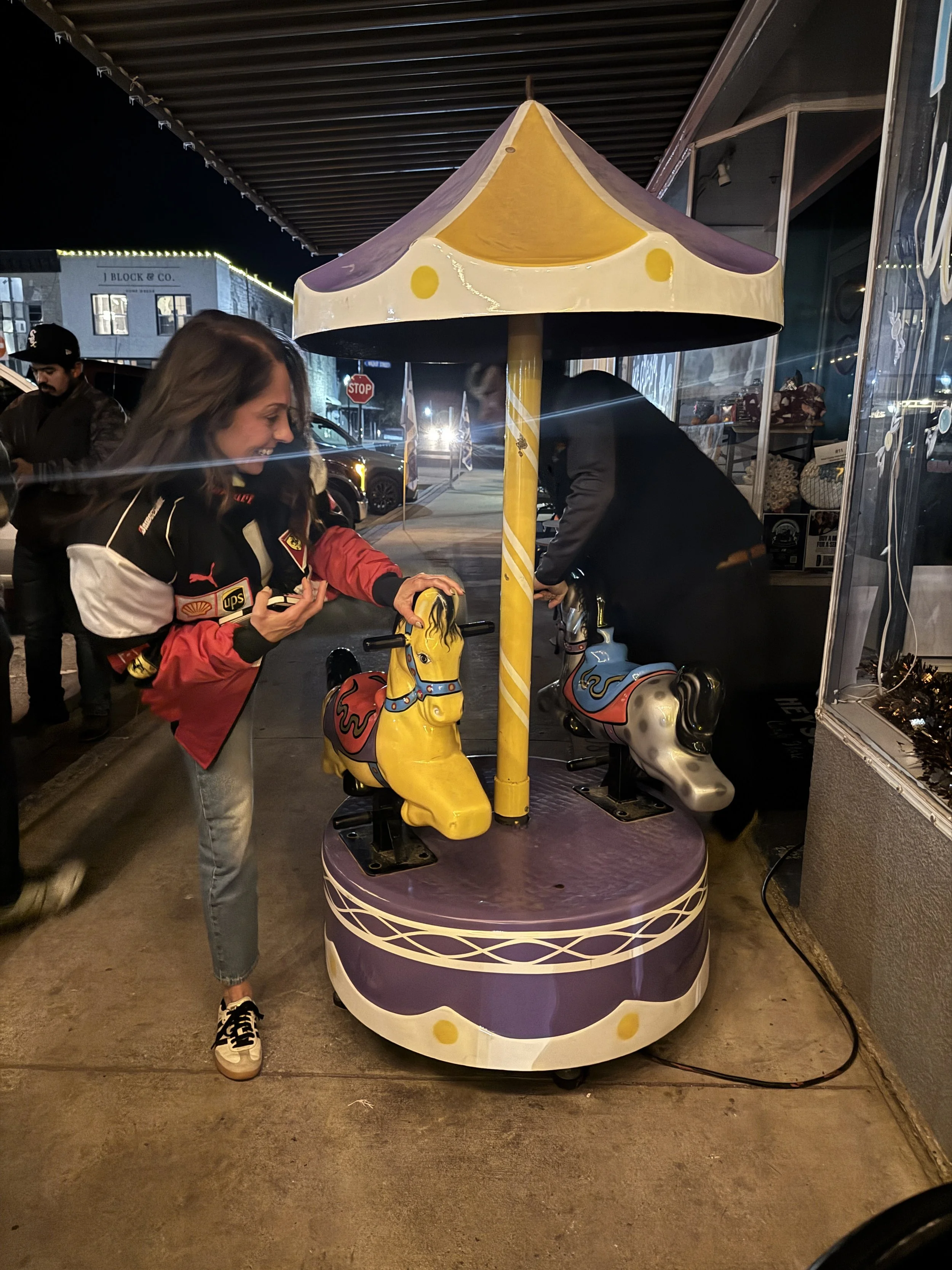 A woman and a man playing on a small, purple carousel with yellow and white horses, underneath a yellow and purple striped canopy, outside at night.
