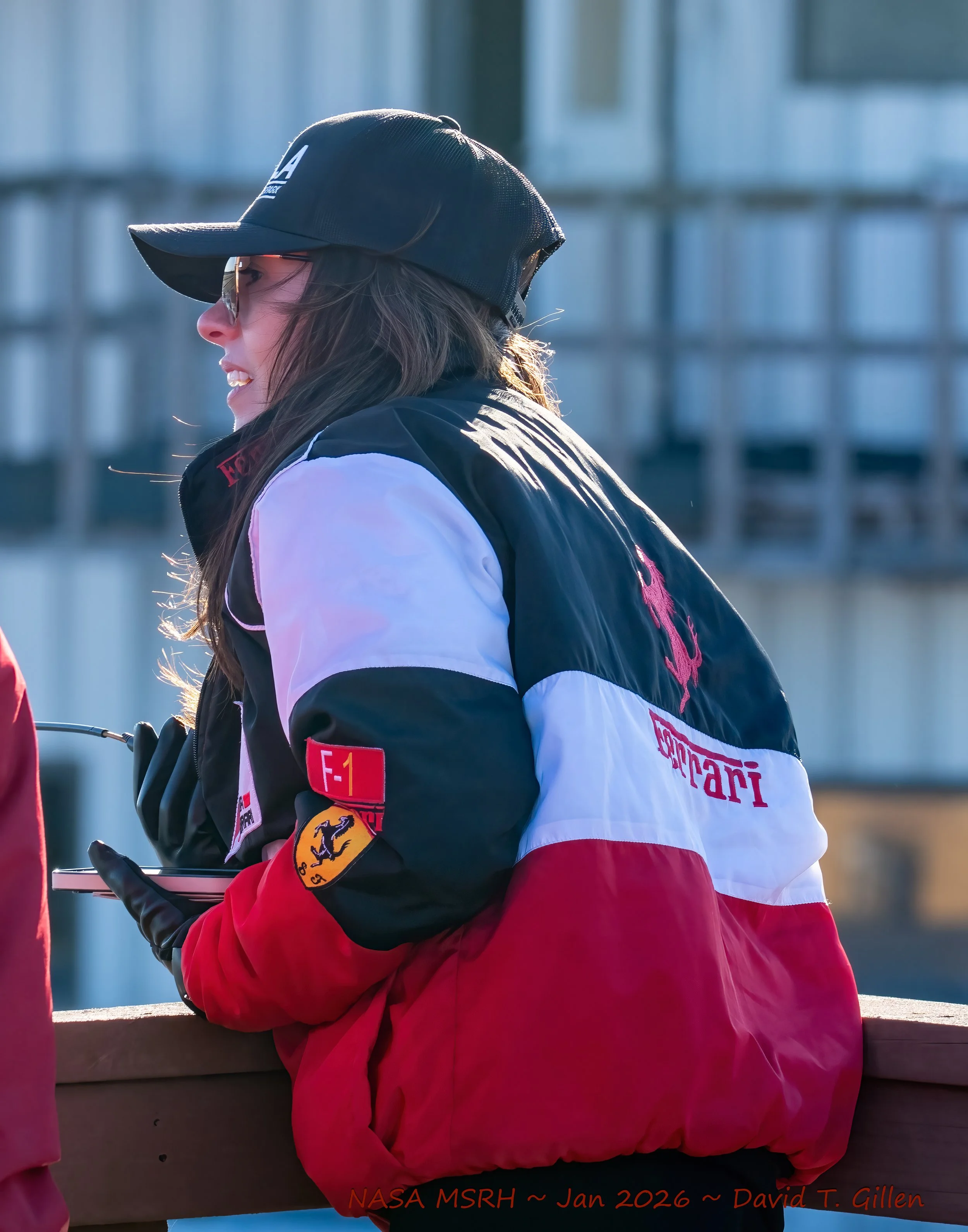 Woman wearing a Ferrari jacket, black cap, sunglasses, and gloves, holding a phone outdoors.