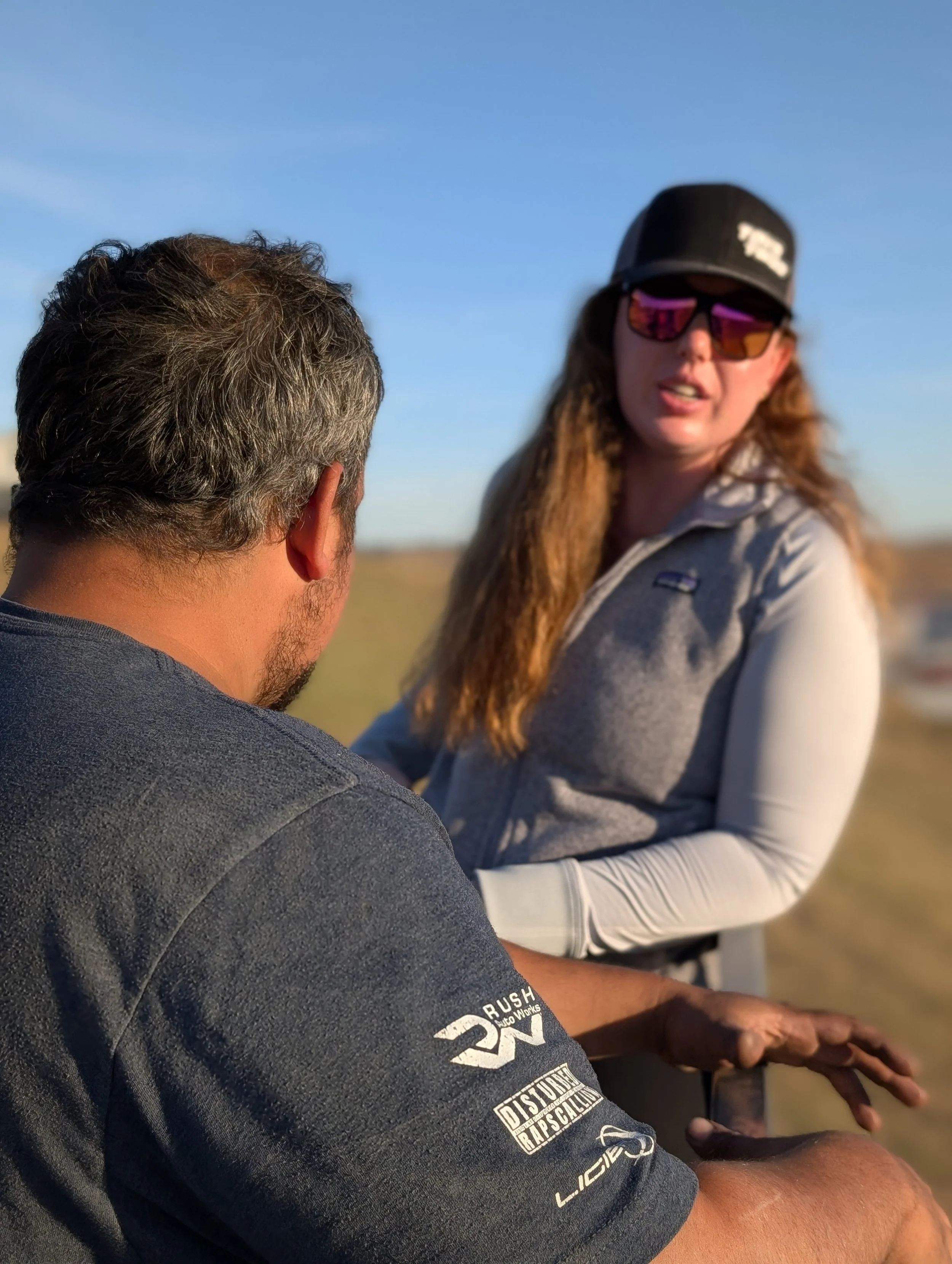A man and a woman conversing outdoors on a sunny day, with the woman wearing sunglasses and a cap, and the man showing the back of his head and shoulder.