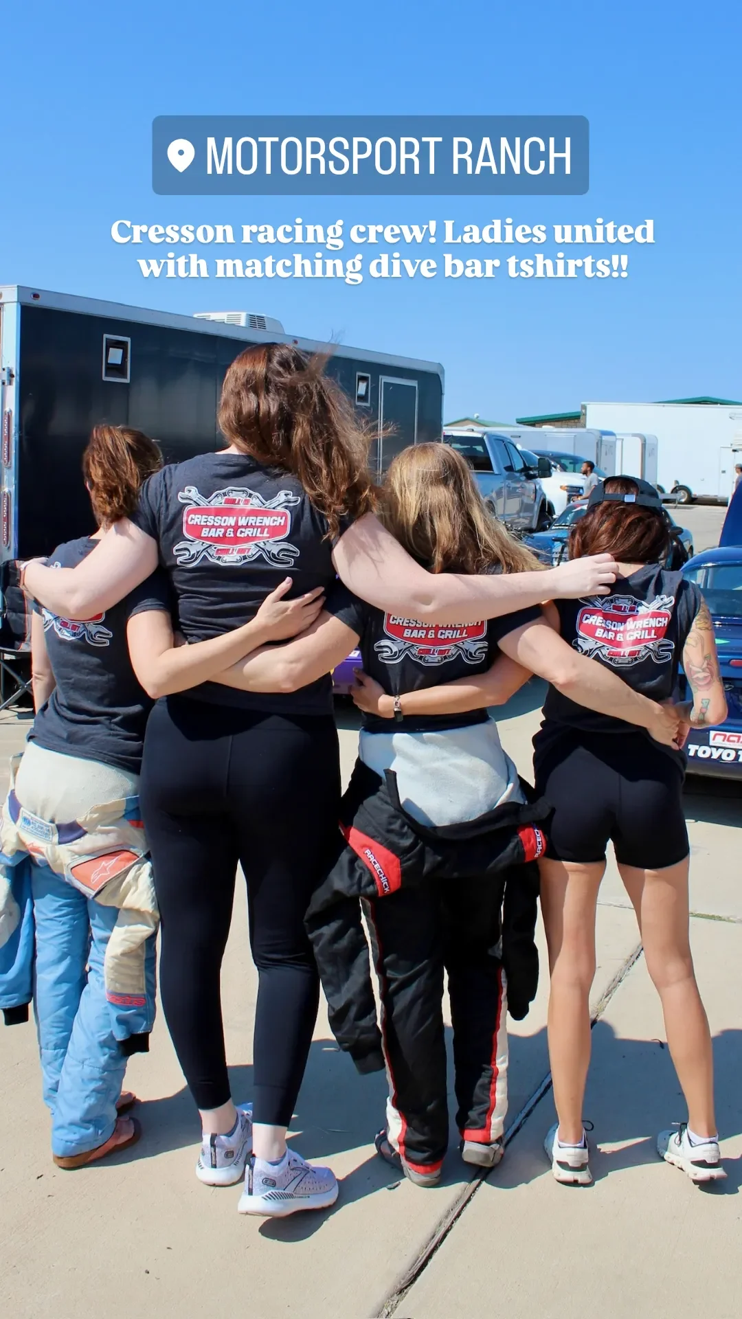A group of women wearing matching black t-shirts with a red and white logo that says 'Cresson Wrench Bar & Grill,' embracing each other in a huddle at the Motorsport Ranch outdoor event with cars and trailers in the background.