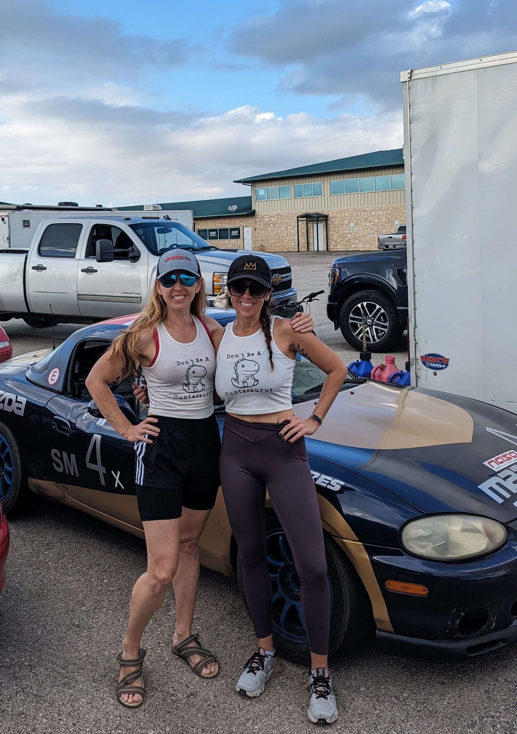 Two women standing in front of a race car at a parking lot, both wearing matching tank tops with a cartoon dinosaur and the words 'Don't Be A Cuntasaurus'. They are smiling, wearing sunglasses and caps, with vehicles and a building in the background.