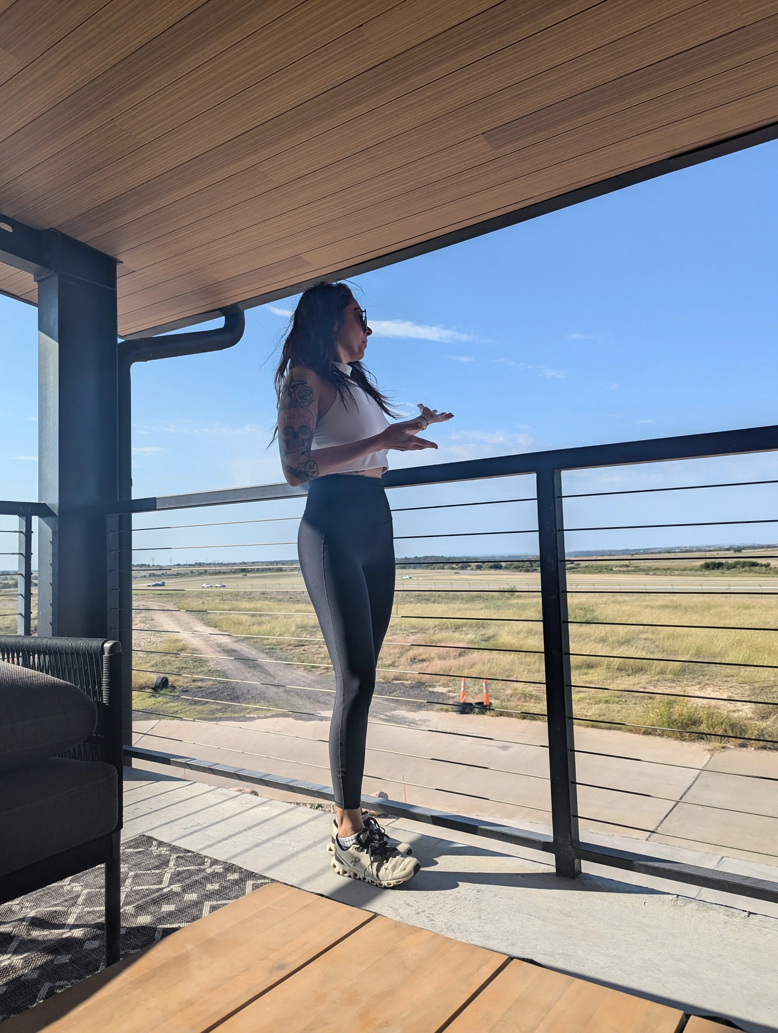 A woman with tattoos wearing workout clothes and sneakers stands on a balcony overlooking a flat open landscape with a clear blue sky.