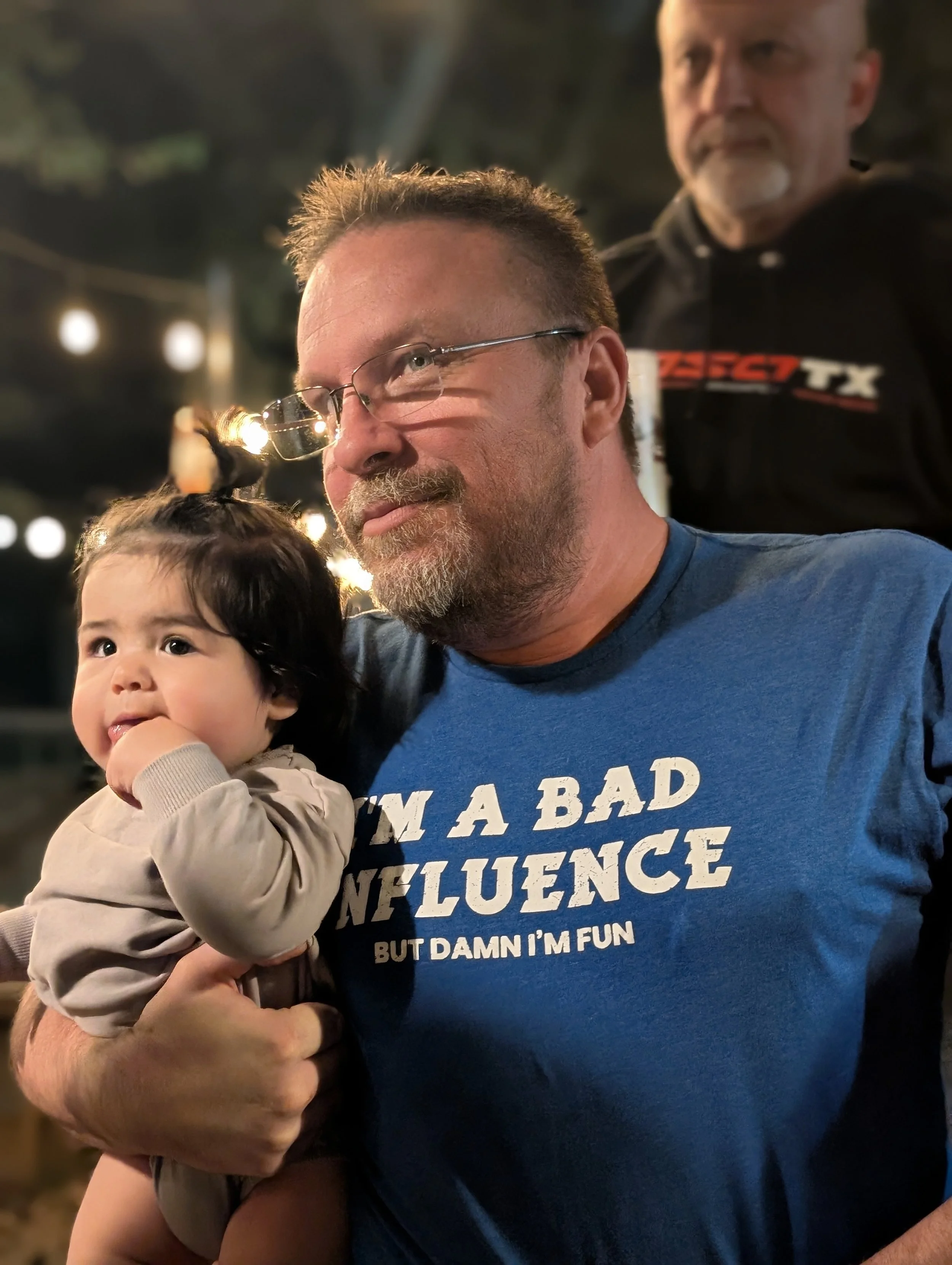 A man holding a young child at an outdoor event during evening. The man is wearing a blue T-shirt with the text "I'M A BAD INFLUENCE BUT DAMN I'M FUN". There is another man with a beard and mustache in the background, wearing a black shirt. The setti