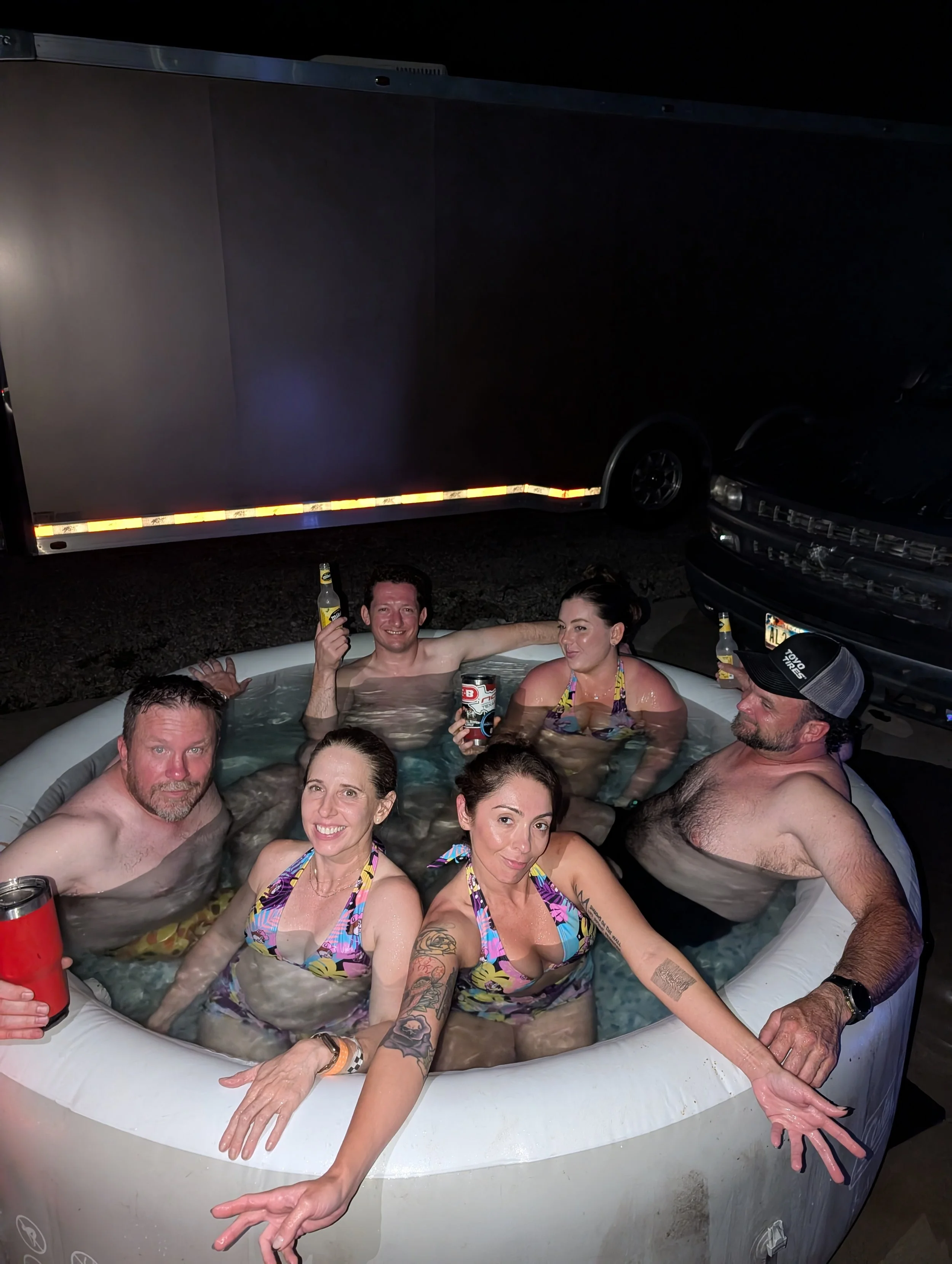 Group of six adults enjoying a hot tub at night, some holding beer bottles, smiling and posing for the photo.