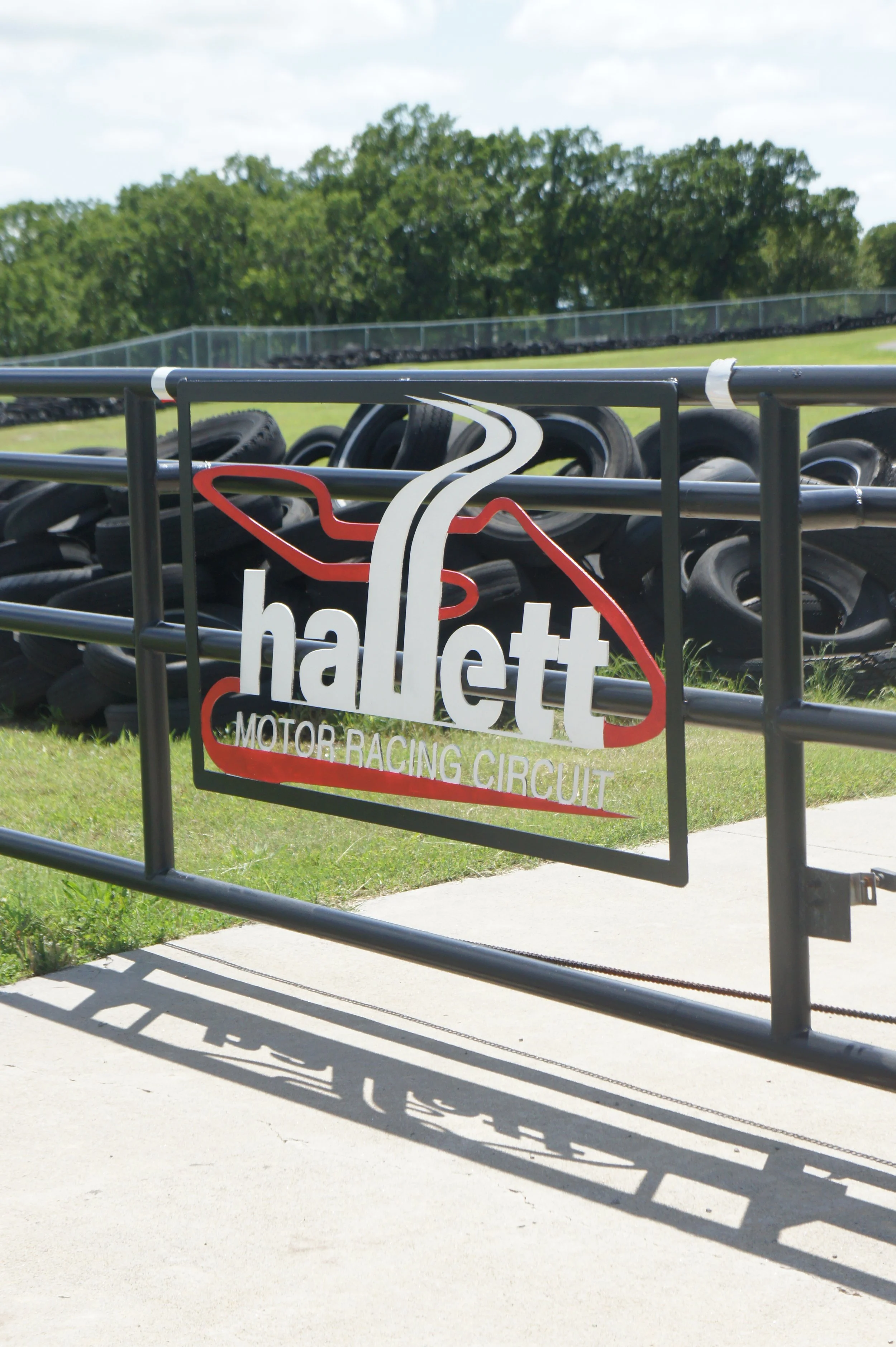 Sign for the Hallett Motor Racing Circuit with racing tires in the background and a grassy area with trees.