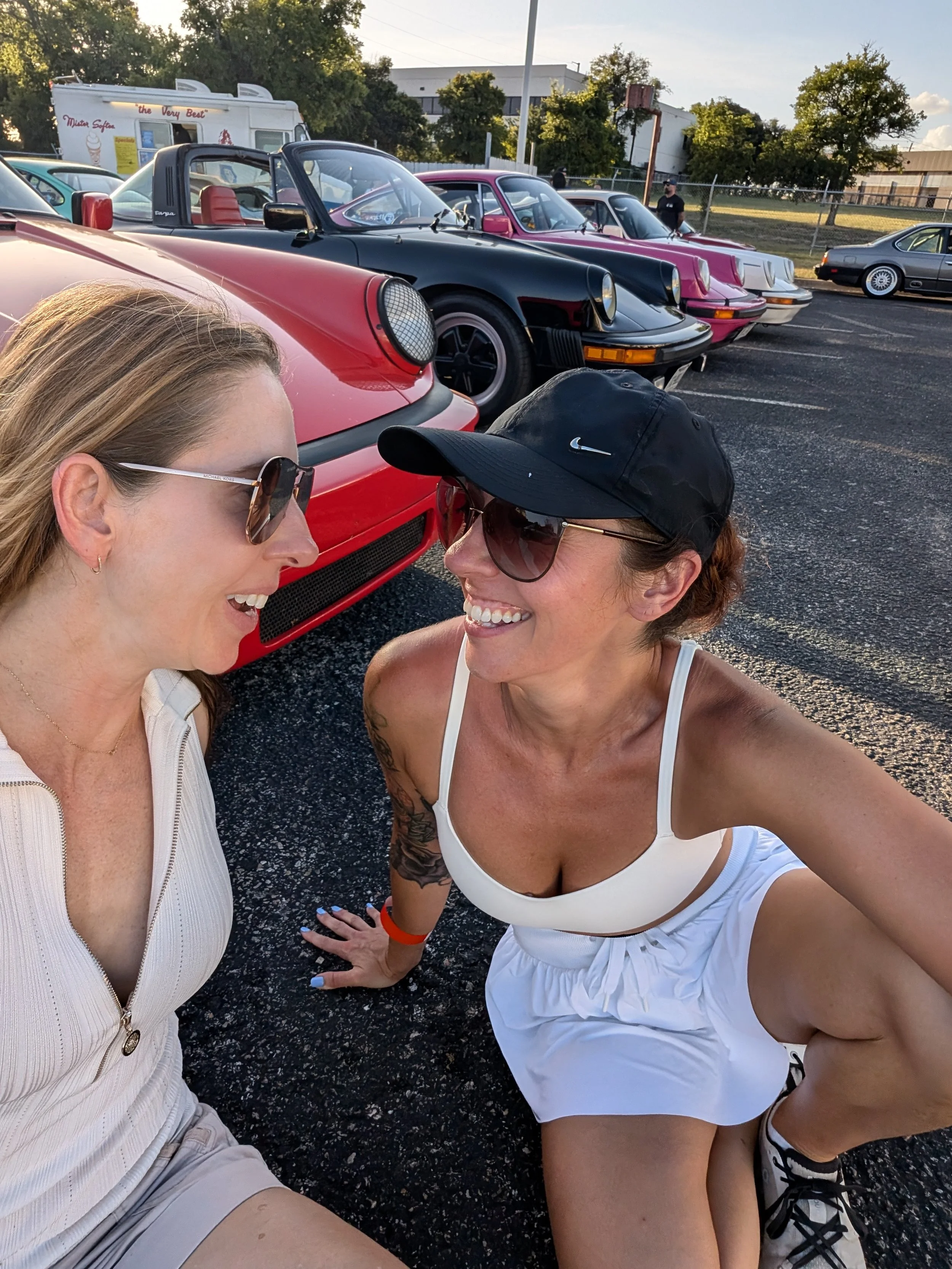 Two women smiling and talking in a parking lot with a row of vintage Porsche cars in the background.