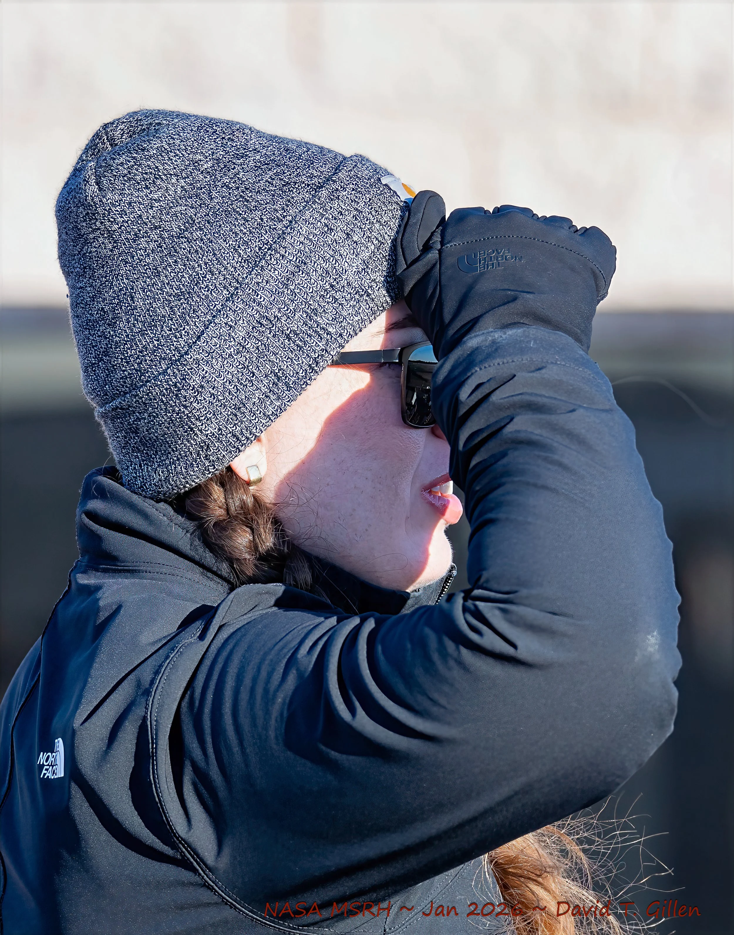 Person wearing a gray beanie, sunglasses, and a black jacket, looking at something in the sky.
