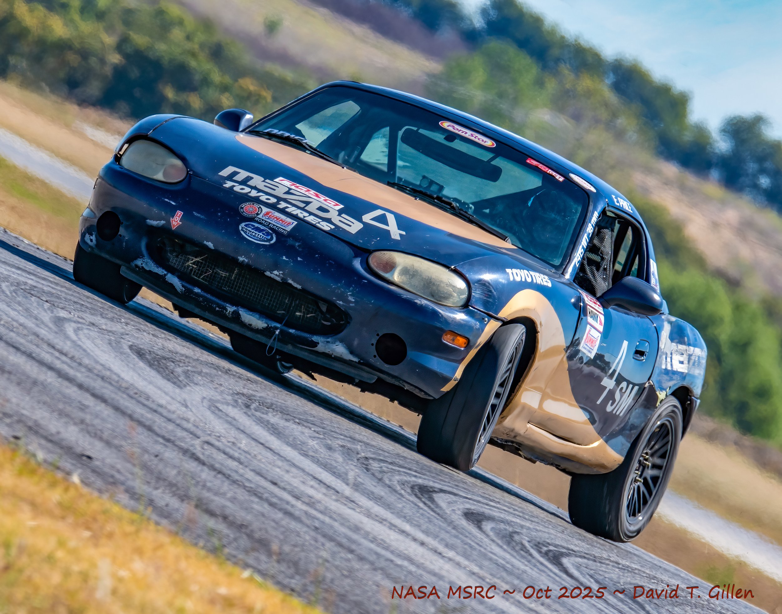 A blue and beige race car with sponsor decals, making a turn on a racetrack, with a slight tilt, green trees, and a blue sky in the background.
