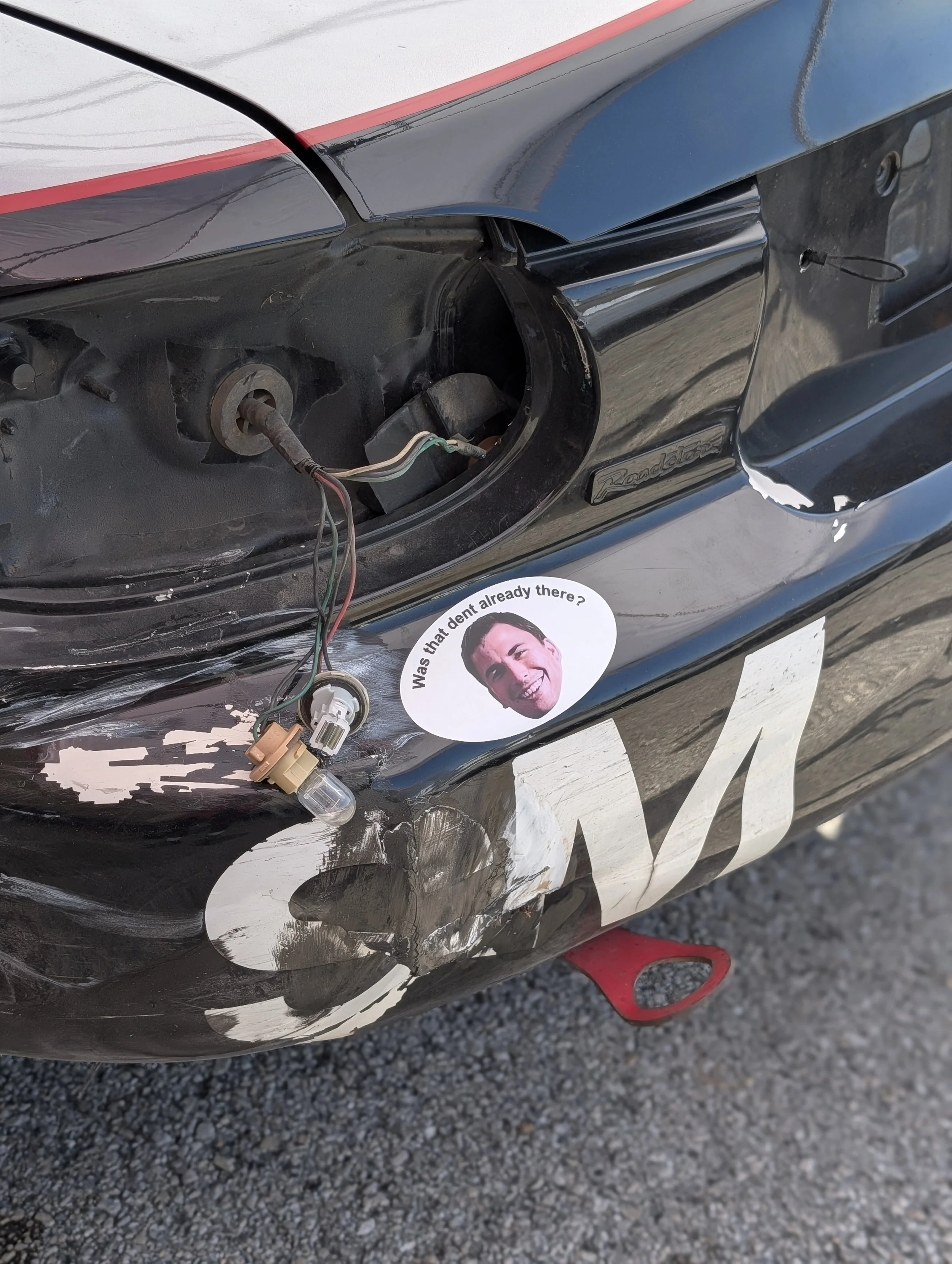 Front of a damaged black race car with a missing headlight, exposed wiring, and a joke sticker showing a smiling man with the caption 'Was that dent already there?'. The car appears to have a minor accident. The car has some painted or damaged areas 