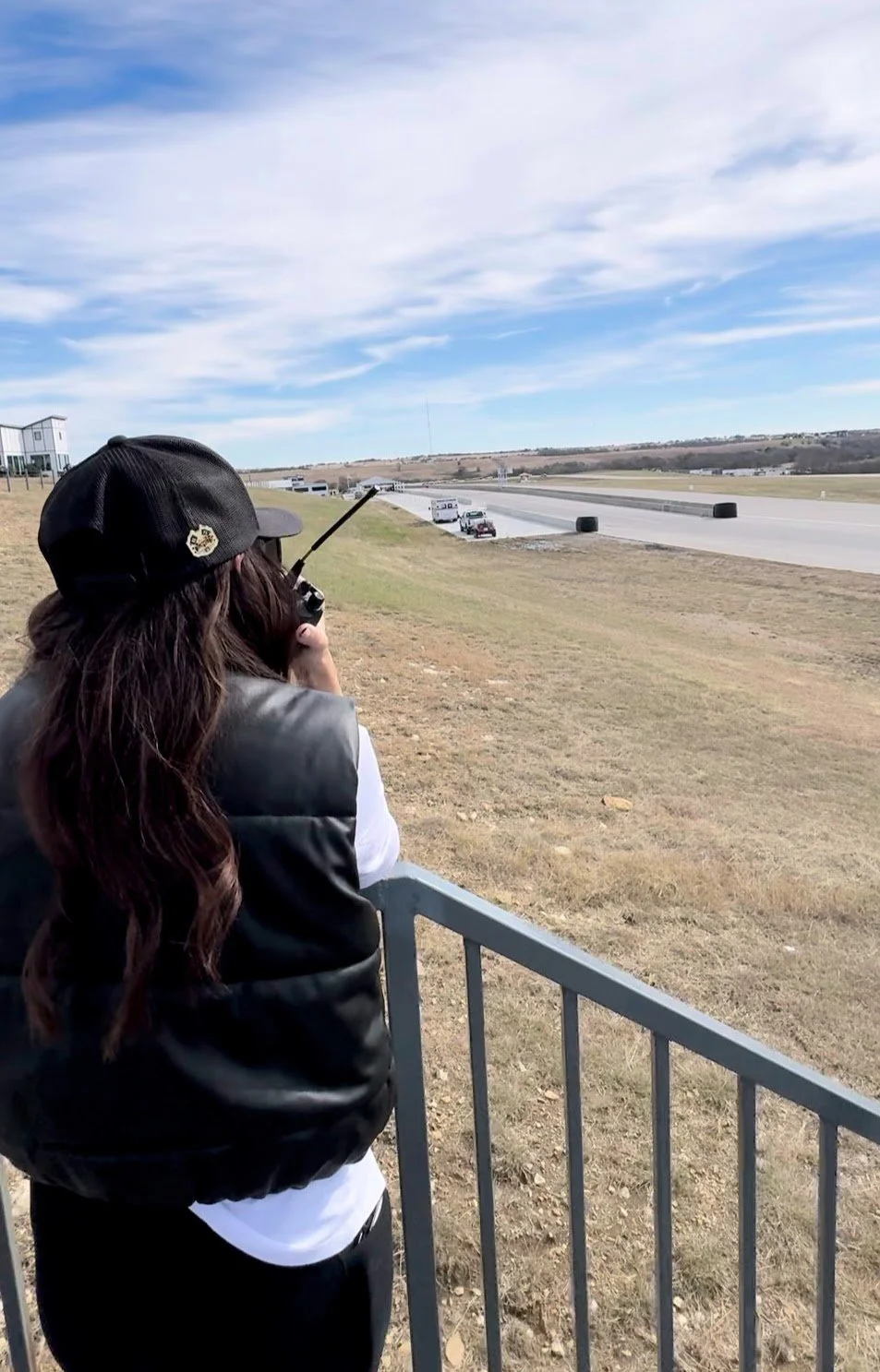 A woman with long brown hair wearing a black cap and vest, standing behind a metal fence, looking at and controlling a remote in her hand, at a race track with cars and trucks on the track and an open landscape with blue sky and clouds in the backgro