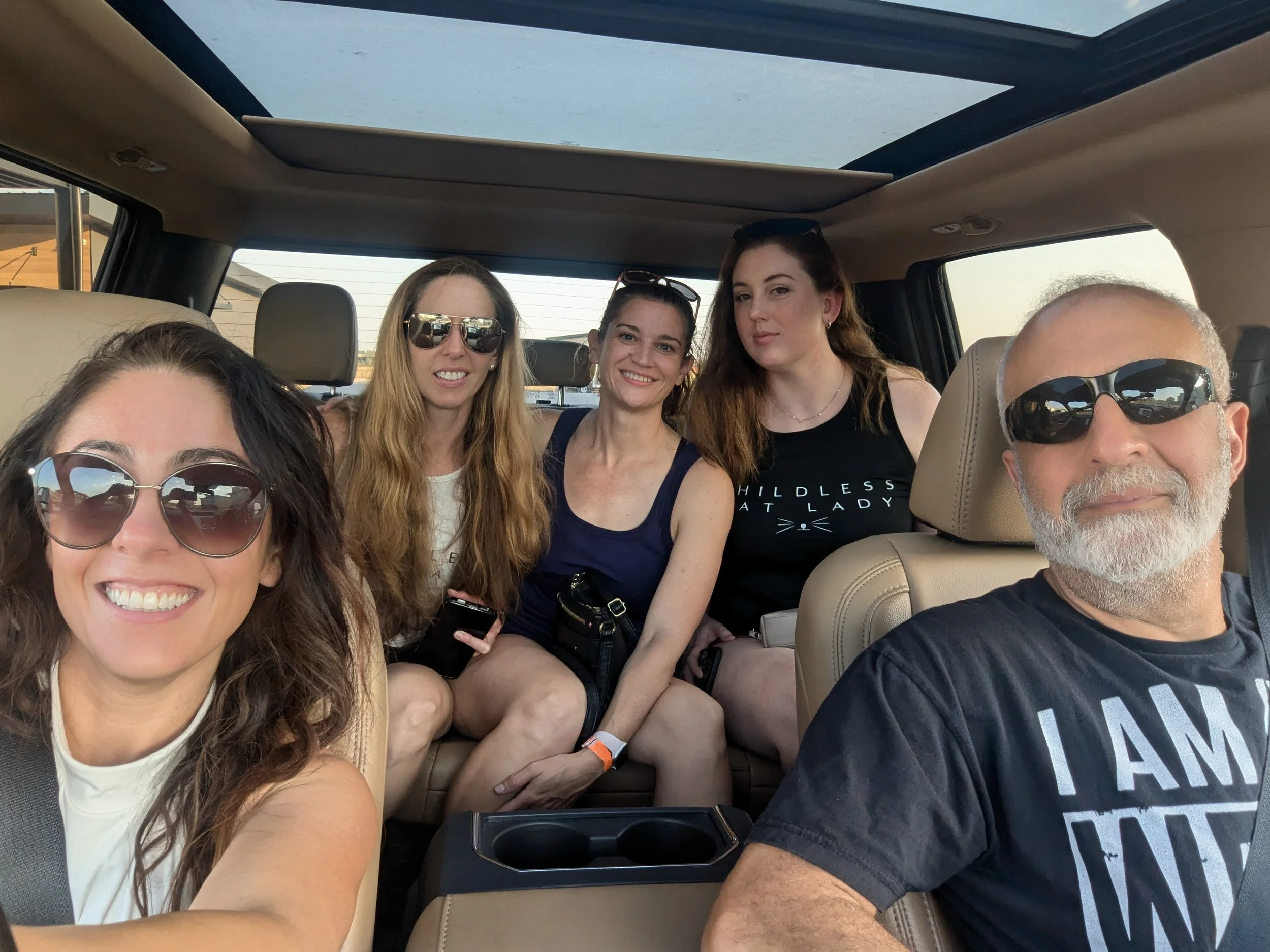 A group of five smiling adults inside a vehicle, four women and one man, wearing sunglasses, taking a selfie on a sunny day.