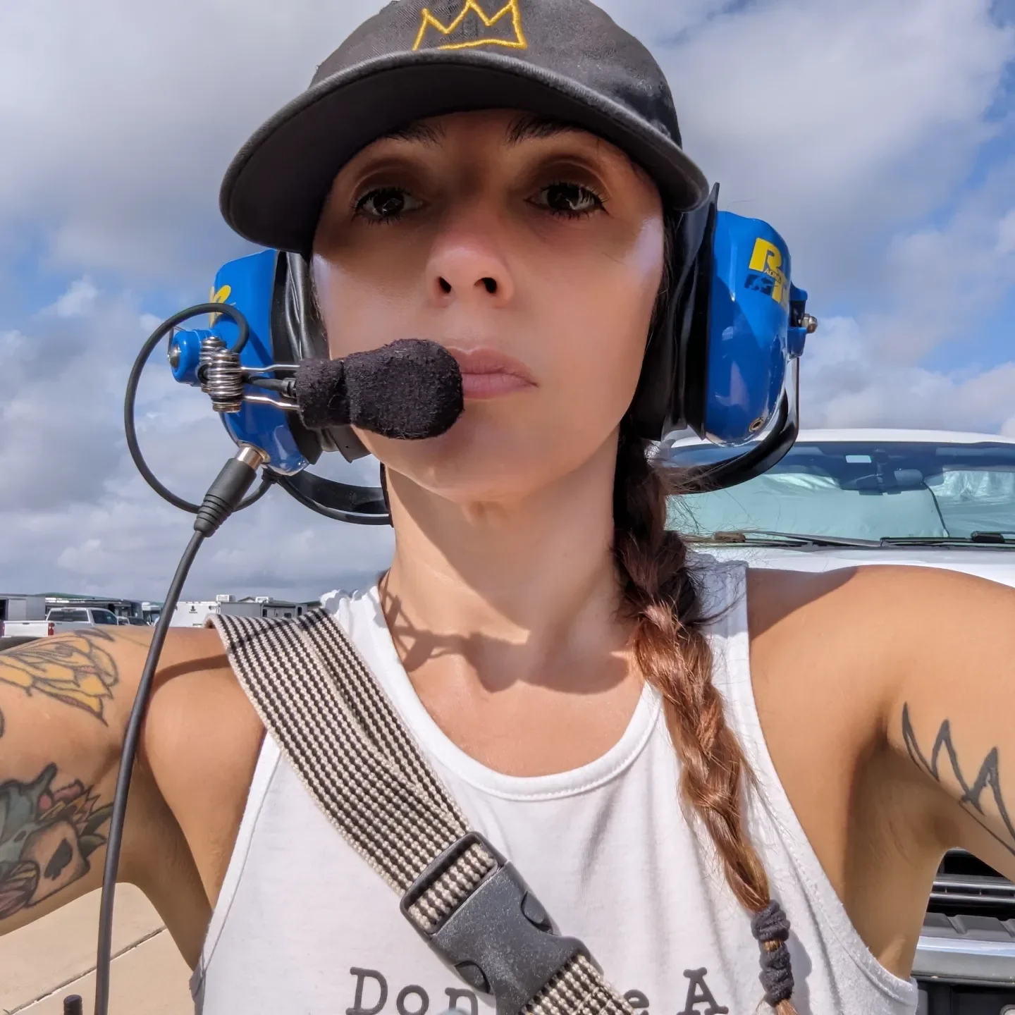 Woman wearing a pilot headset and cap, taking selfie outdoors on a cloudy day.