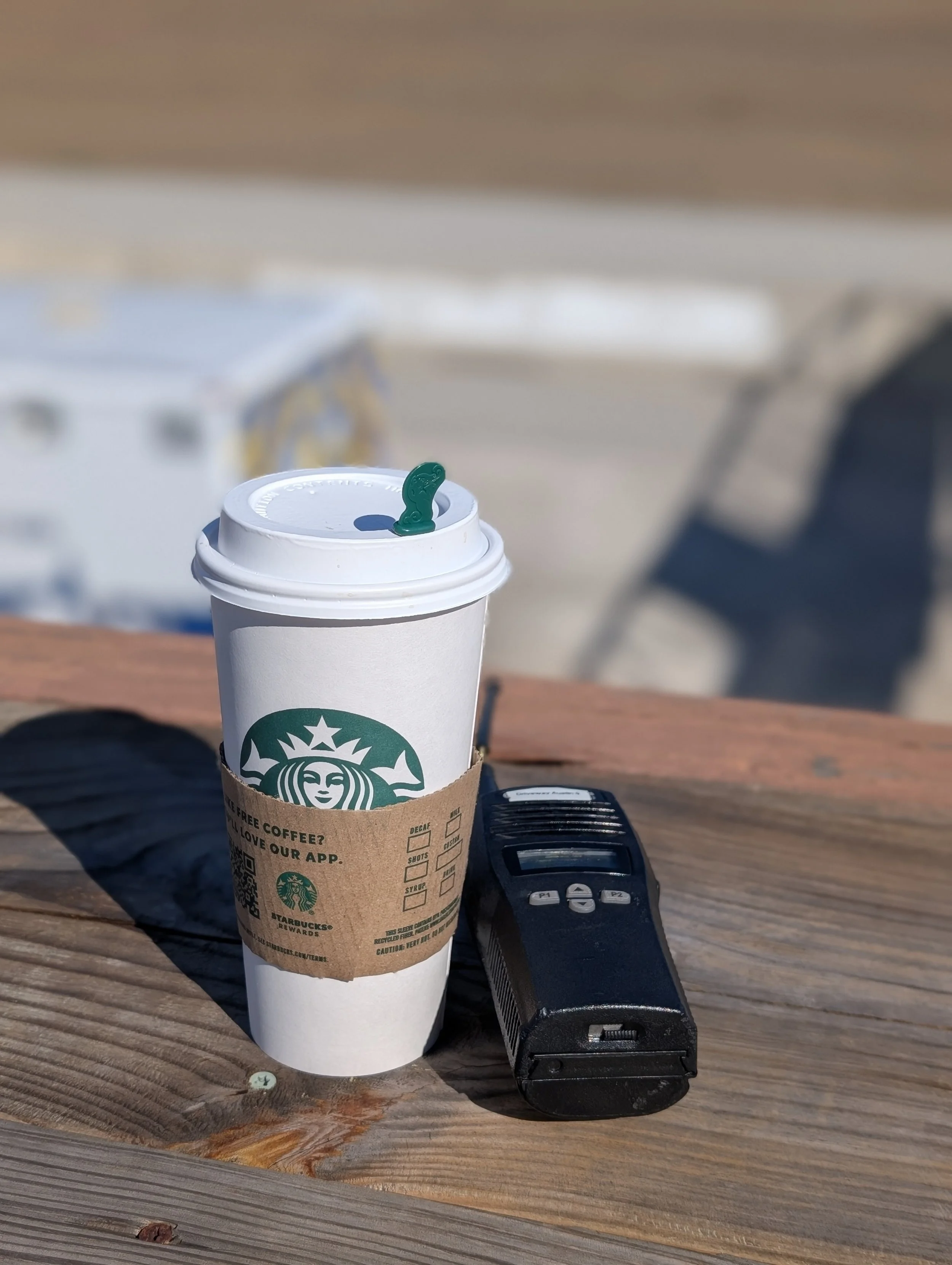 A to-go Starbucks coffee cup with a green straw, a brown sleeve, and a black remote control placed on a wooden surface outdoors, with blurry white and dark objects in the background.