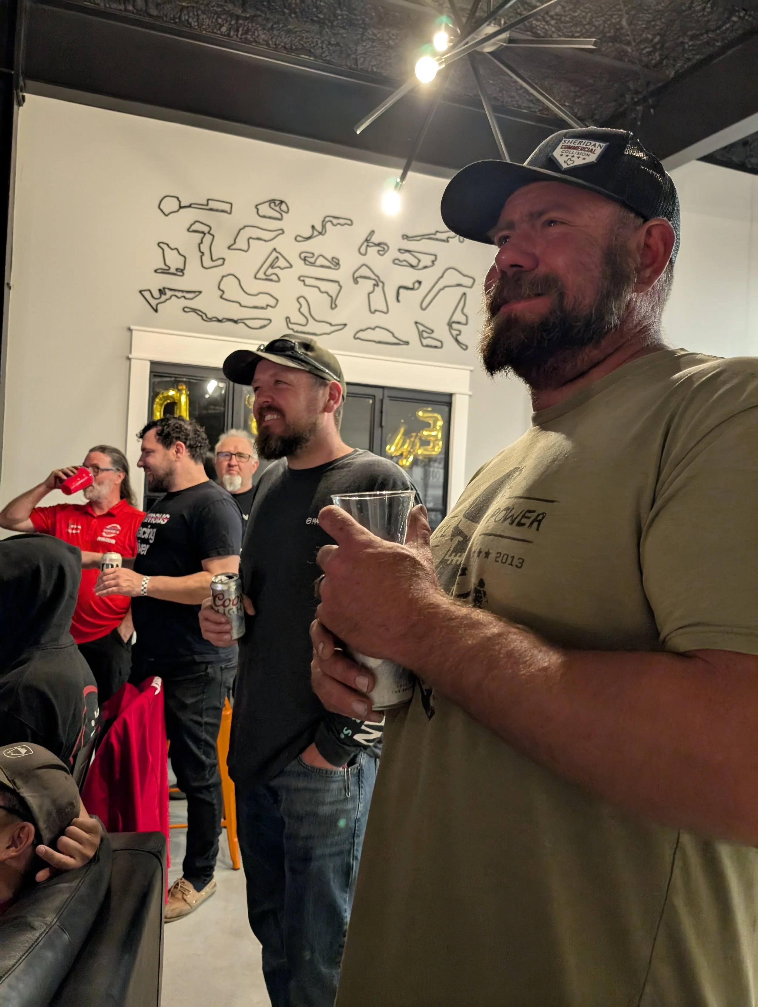 Group of men at a social gathering, some holding drinks and smiling, in a room with modern decor and abstract art on the wall.