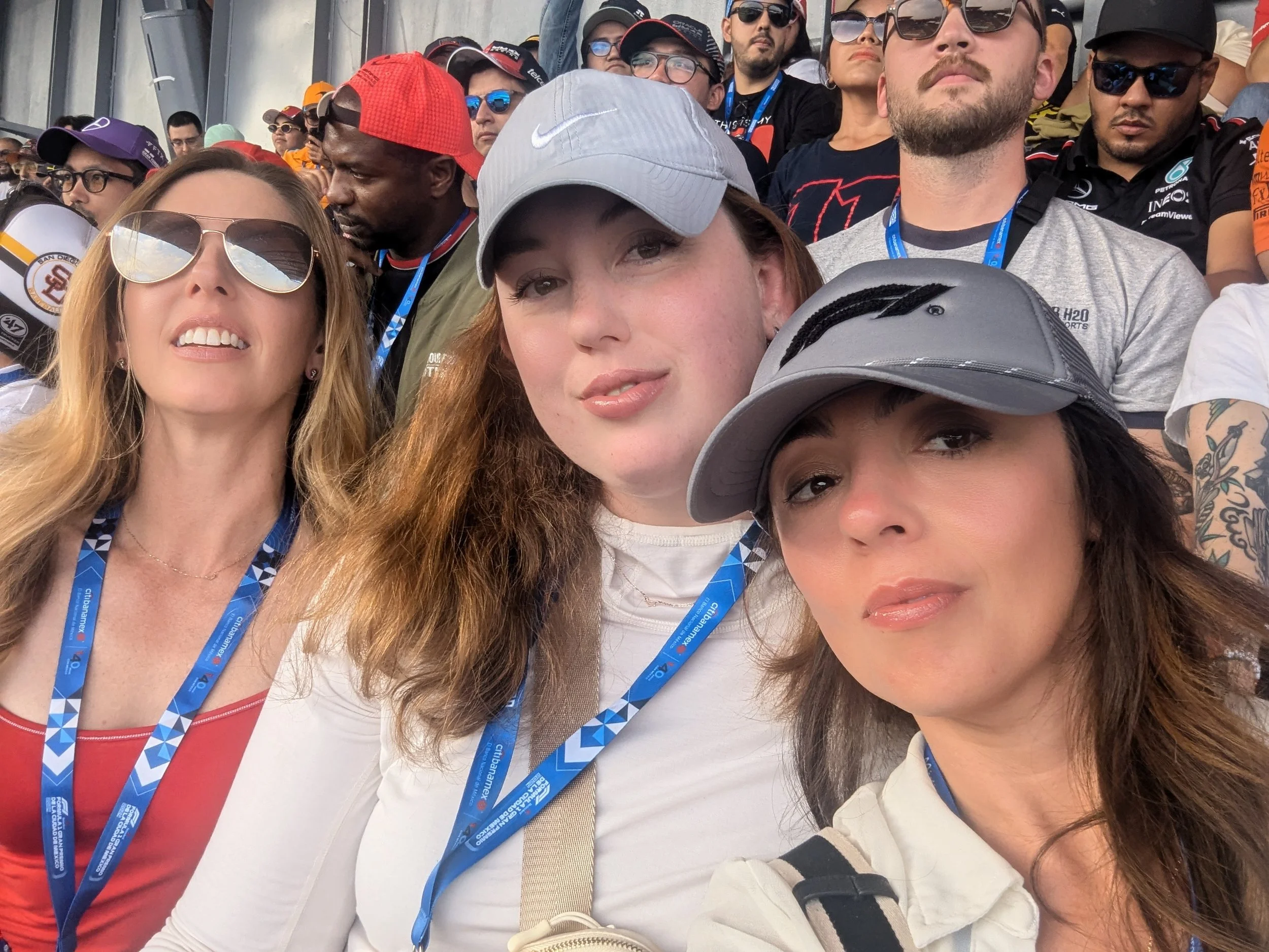 Group of people, including three women in the foreground wearing caps and sunglasses, at a public event or gathering.