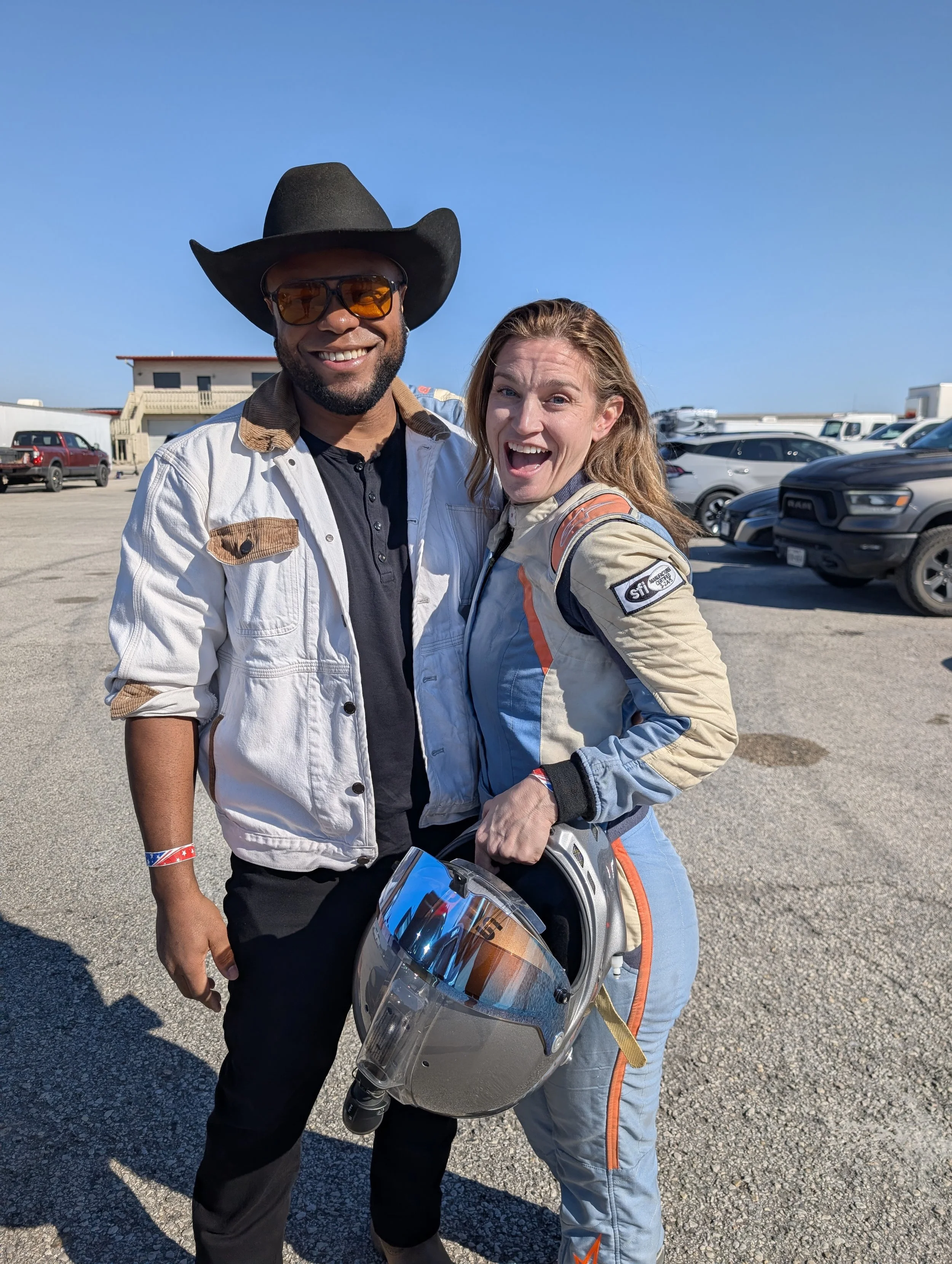Two smiling people, one man wearing a cowboy hat, sunglasses, and a casual jacket, and a woman in a motorcycle racing suit holding a helmet, standing outdoors in a parking lot with cars and a building in the background, under a clear blue sky.