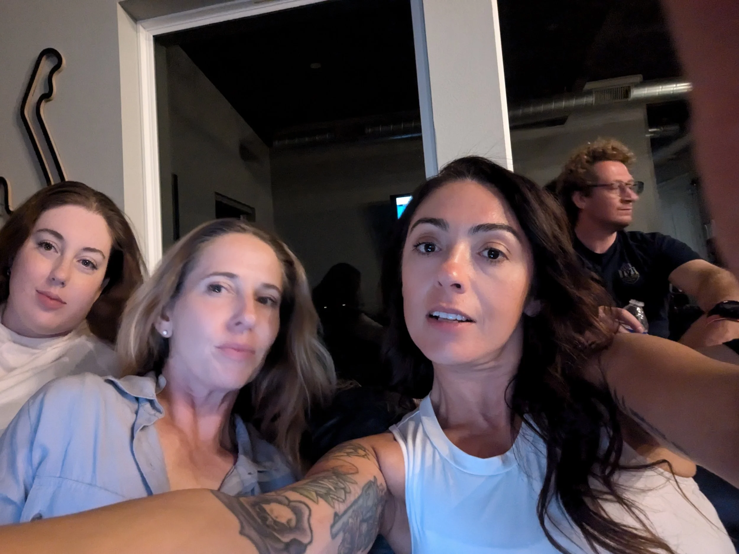 Four people sitting closely together in an indoor setting, taking a selfie. The woman in front has dark hair, a tattoo on her arm, and is wearing a white sleeveless top. The woman to her left has blonde hair and is wearing a gray button-up shirt. The