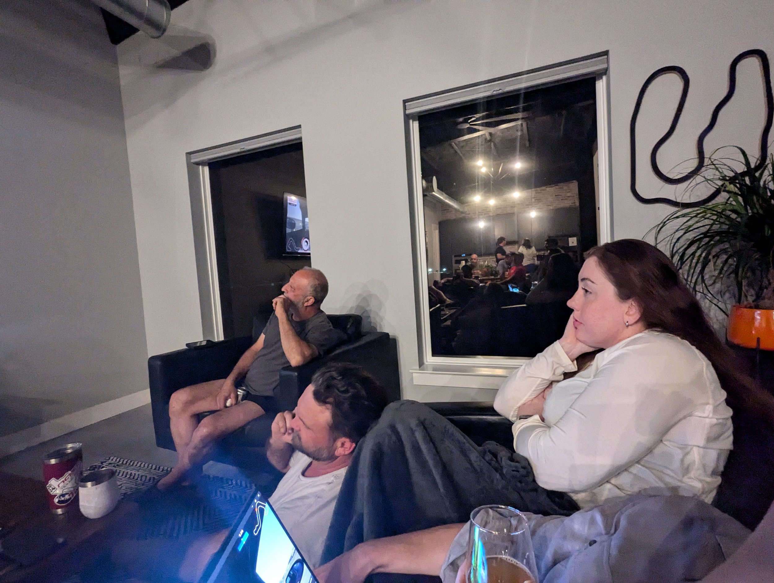 People sitting on couches and chairs in a living room, watching television, with drinks on the table, during evening or night.