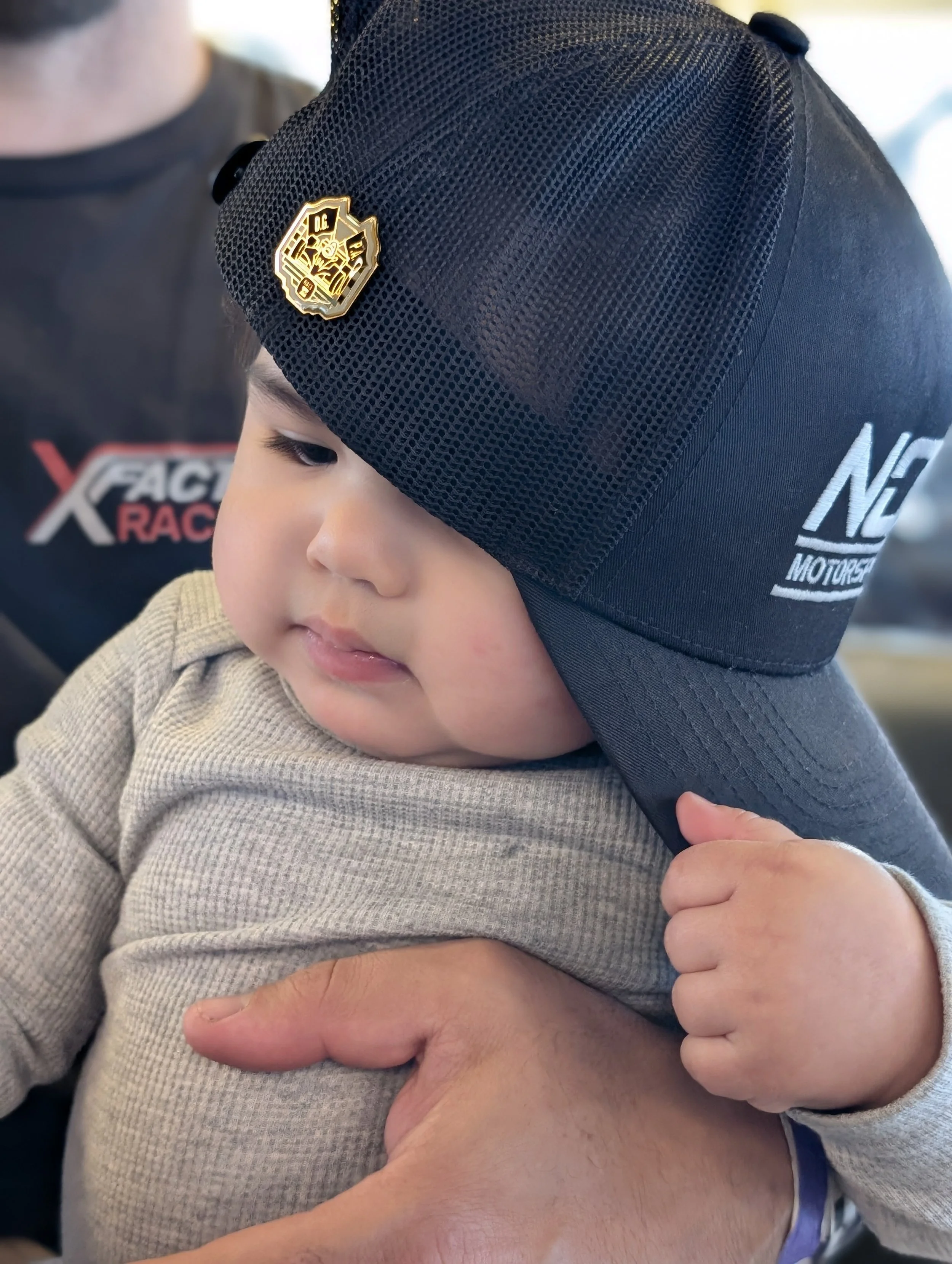 A young child with chubby cheeks wearing a beige long-sleeve shirt and a black mesh cap with race team logos, being held by an adult.