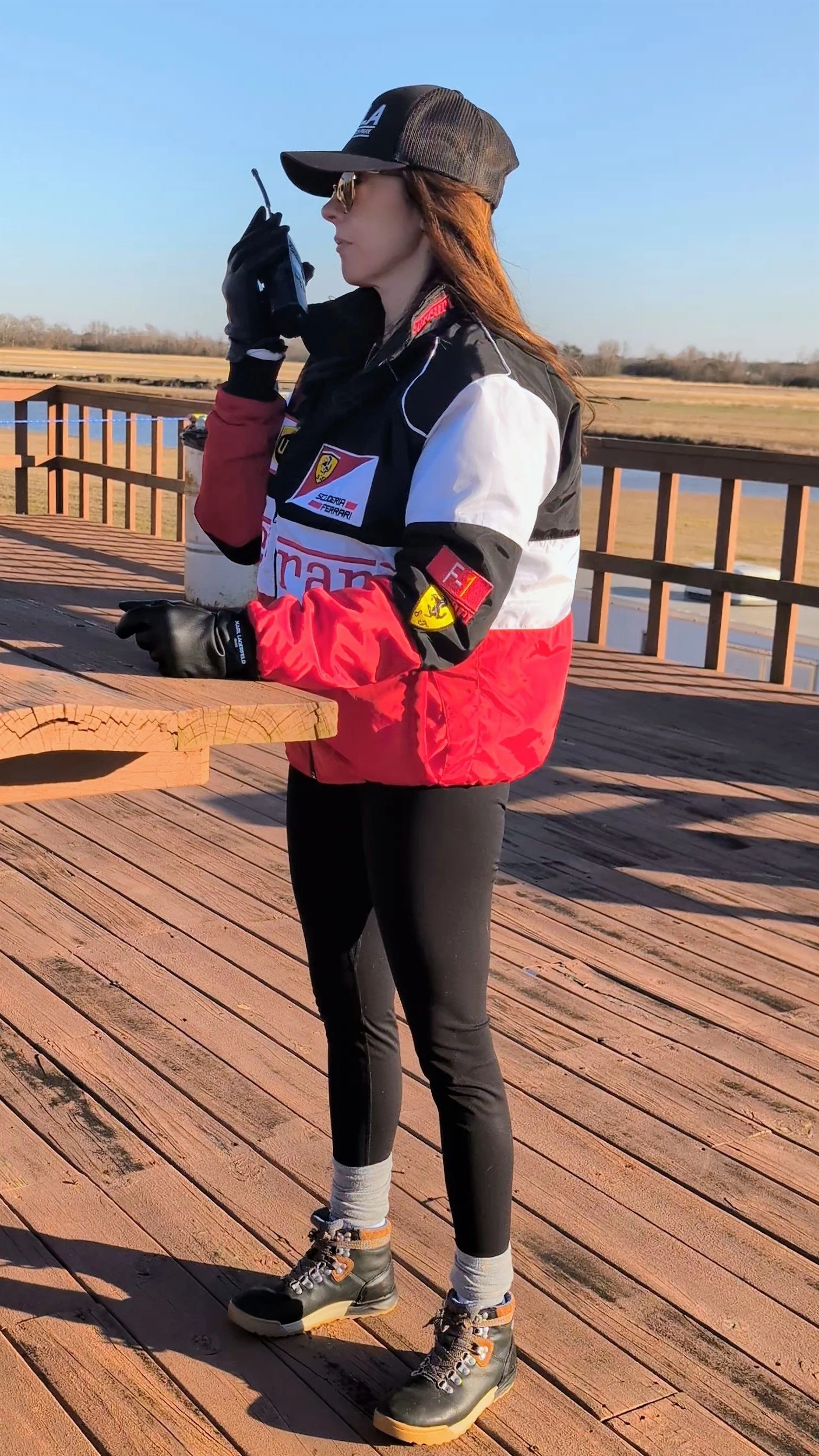 A woman standing on a wooden deck, dressed in racing gear, holding a walkie-talkie, with a scenic landscape of fields and a small body of water in the background.