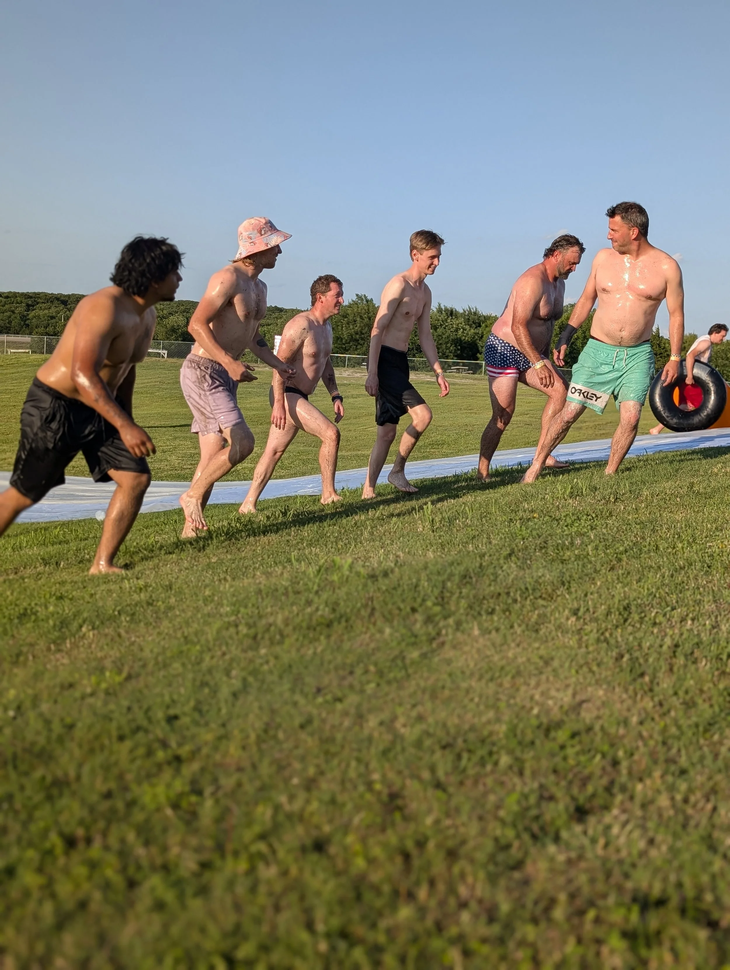 Group of men in swim trunks running on a grassy field, participating in a water-based activity, some holding flotation devices, on a sunny day.
