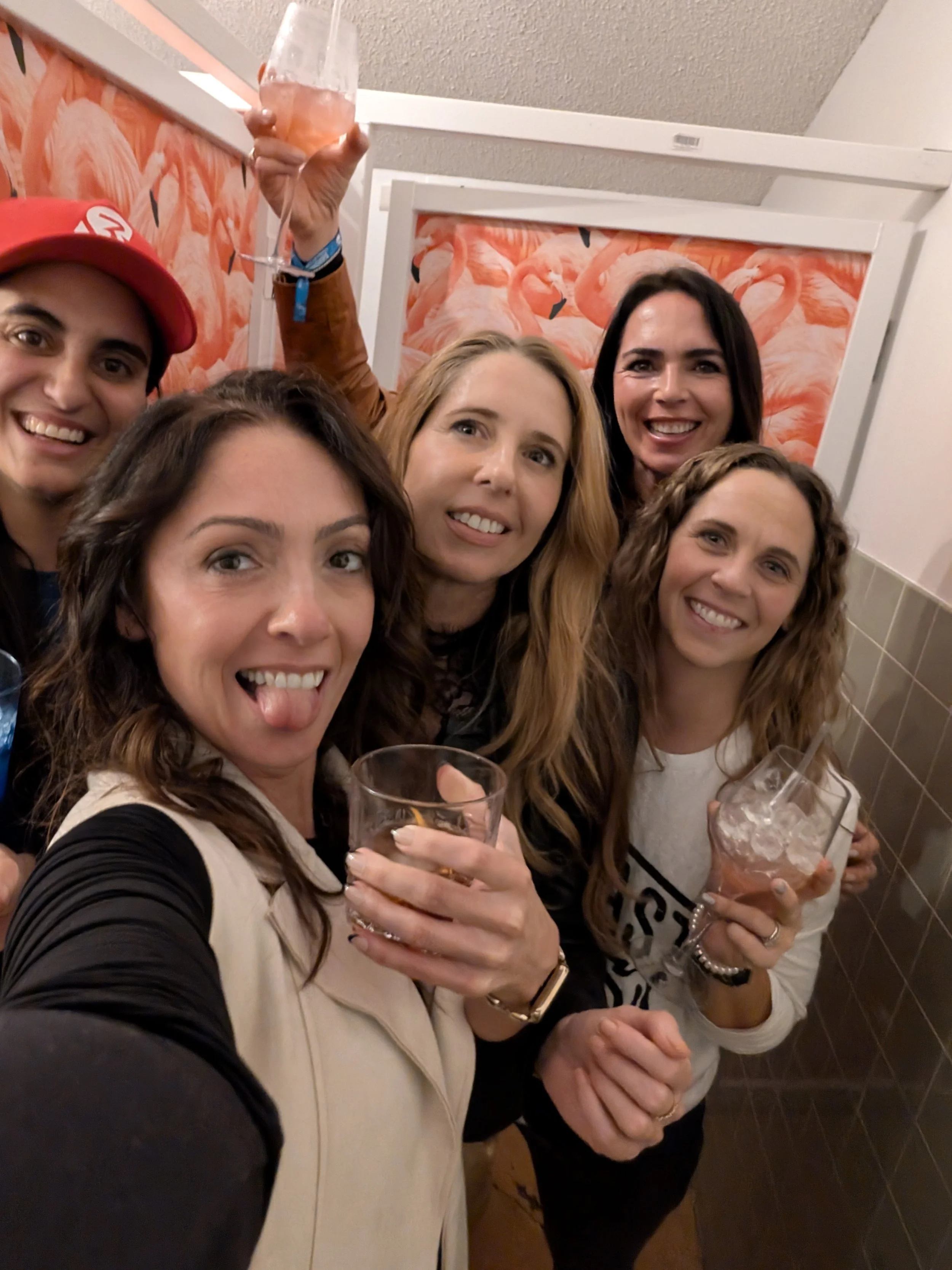 Group of five women celebrating with drinks, smiling, in a room with flamingo-themed wallpaper.