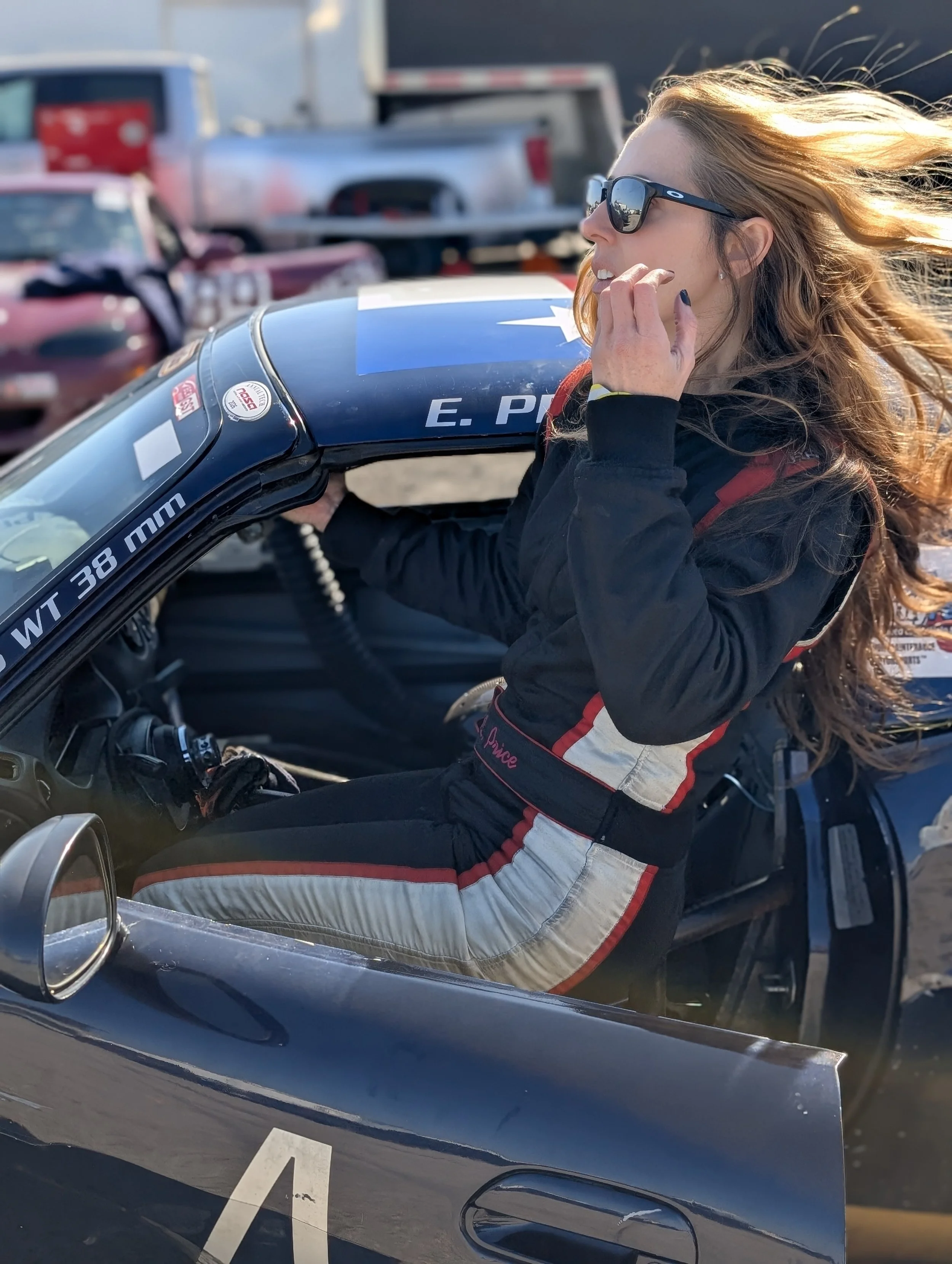 A woman with long red hair and sunglasses sitting in a race car, holding the car's roof, with her hair blowing in the wind and surrounded by parked cars.