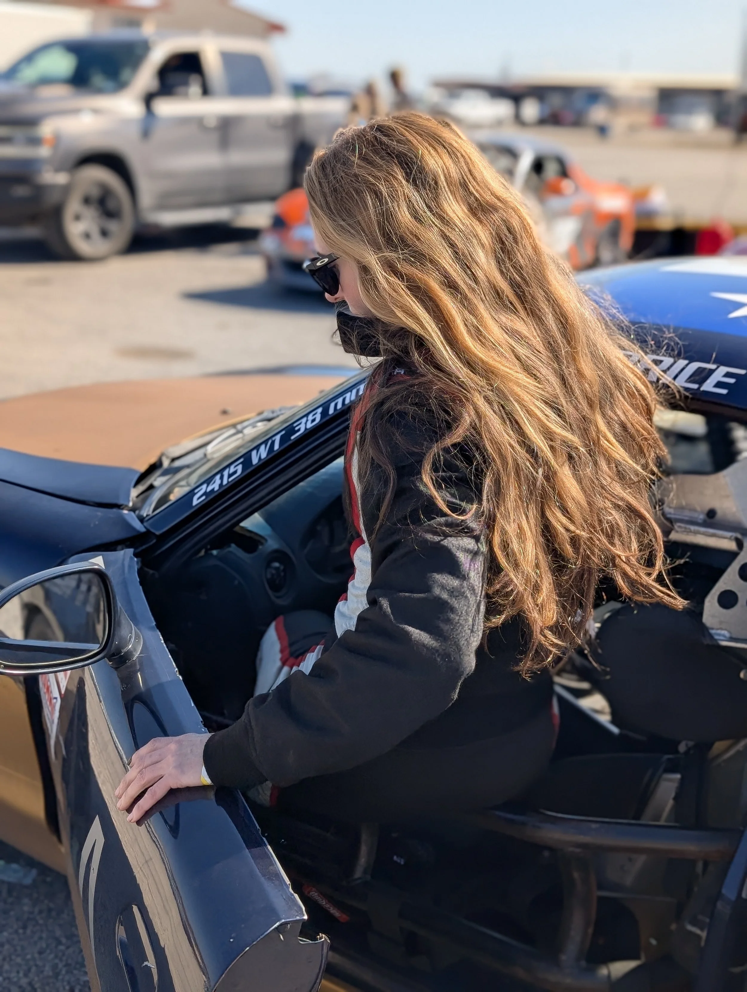 A woman with long, wavy, reddish-blonde hair wearing sunglasses and a racing suit sitting in a race car at a racetrack.
