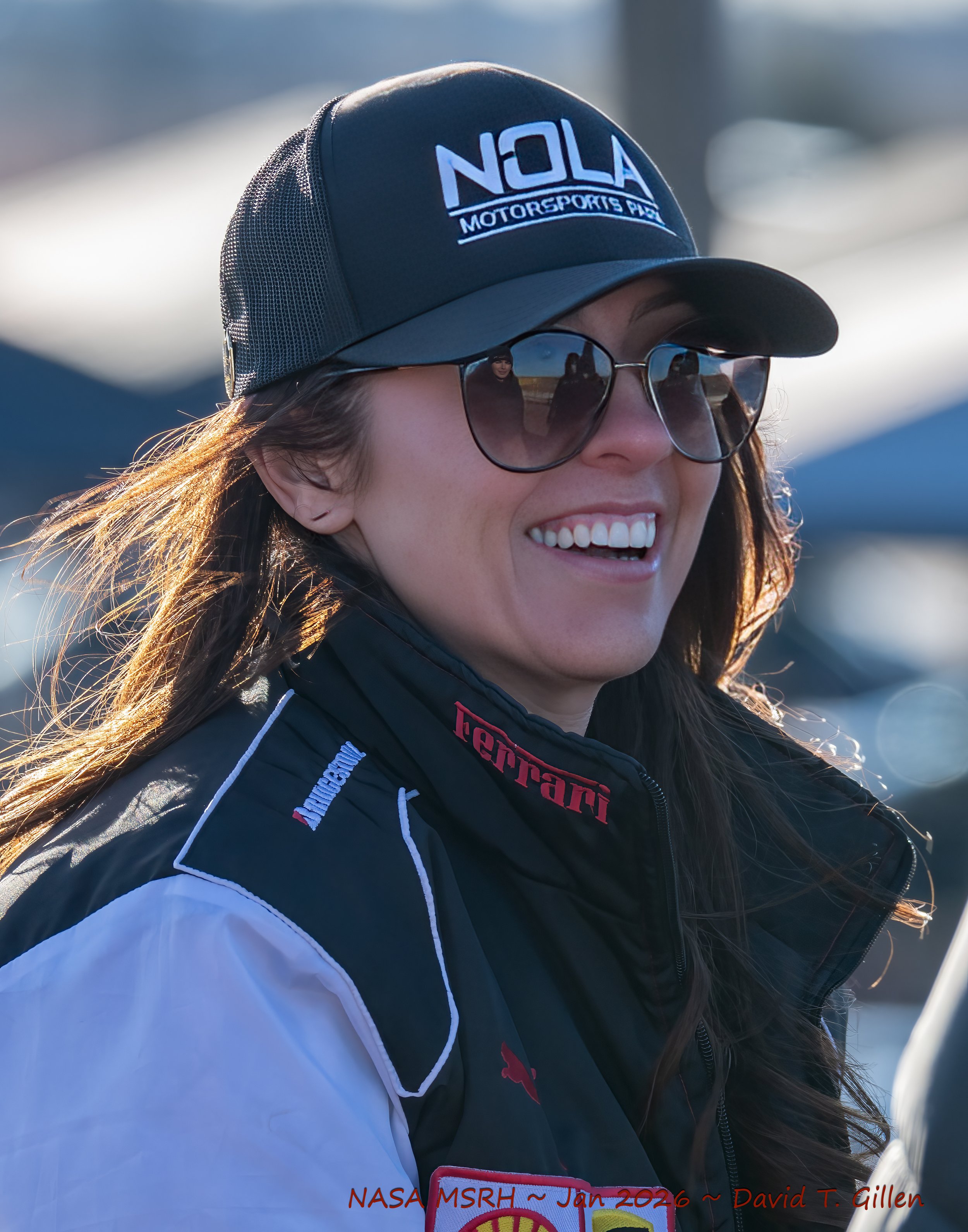 Woman wearing a black cap with 'NOLA Motorsports Park' logo, sunglasses, and racing jacket smiling outdoors with blurred background.