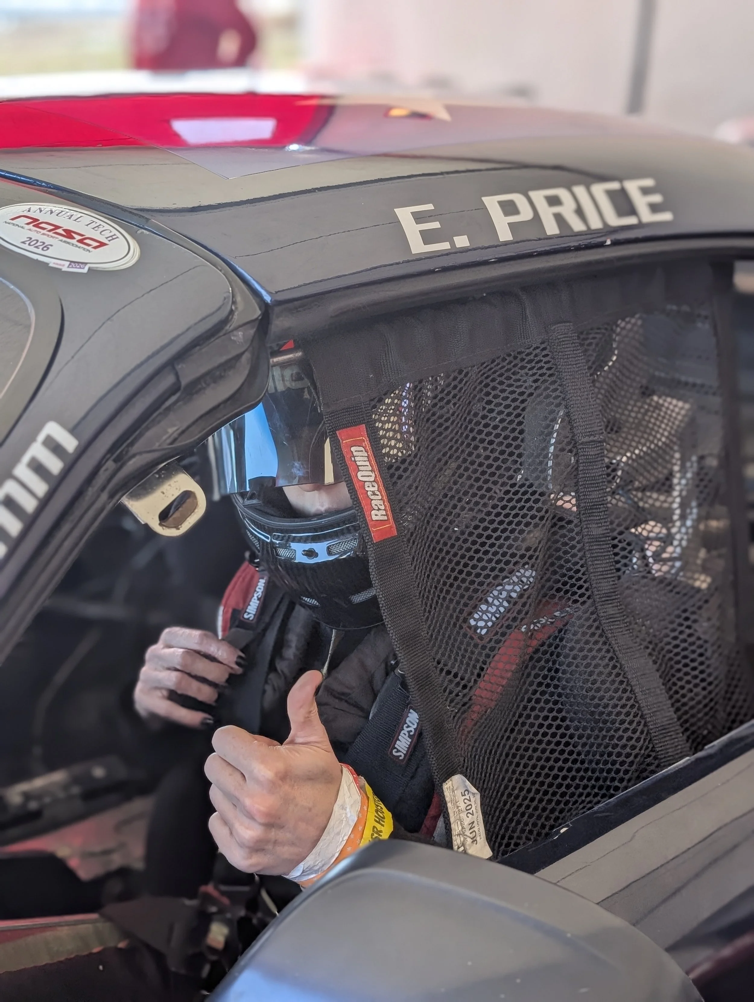 Race car driver wearing a helmet and racing suit sitting inside a race car with a mesh window, showing a thumb up gesture. The car has the name E. Price on the top.