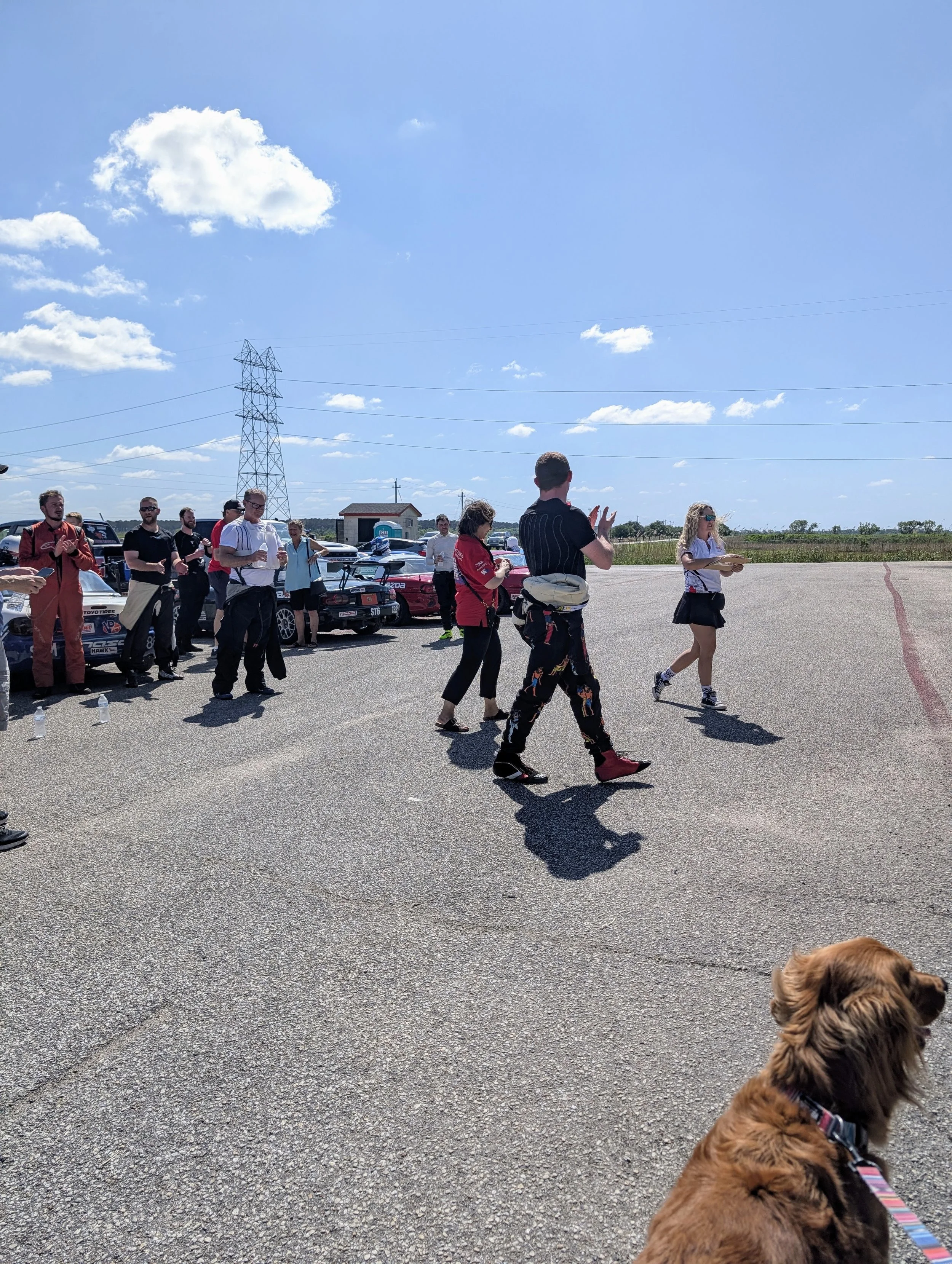 People gathered in an outdoor parking lot, some taking photos and others walking, with cars parked in the background and a clear blue sky with scattered clouds.