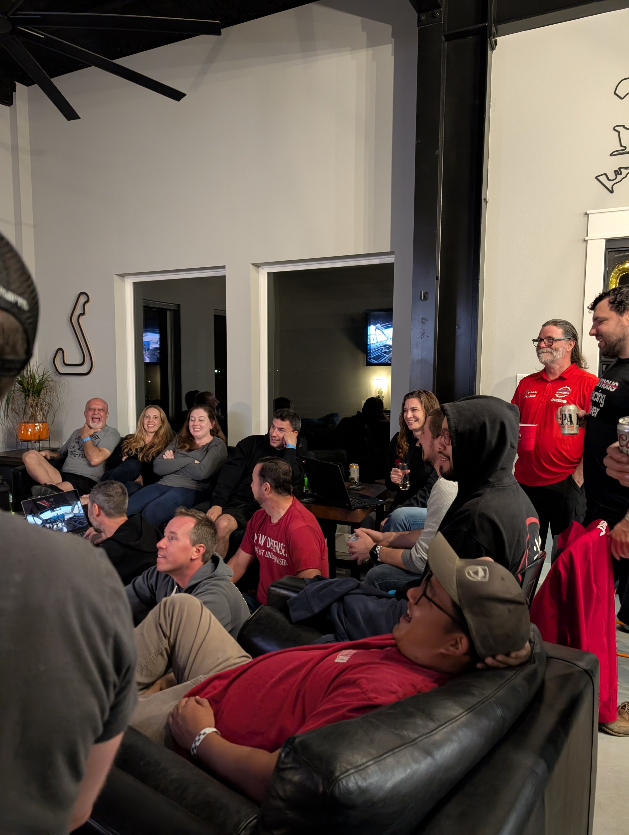 People seated and standing in a living room, watching a sports event on TV, smiling and laughing, with some holding drinks, while a man in a red racing team shirt stands on the right.