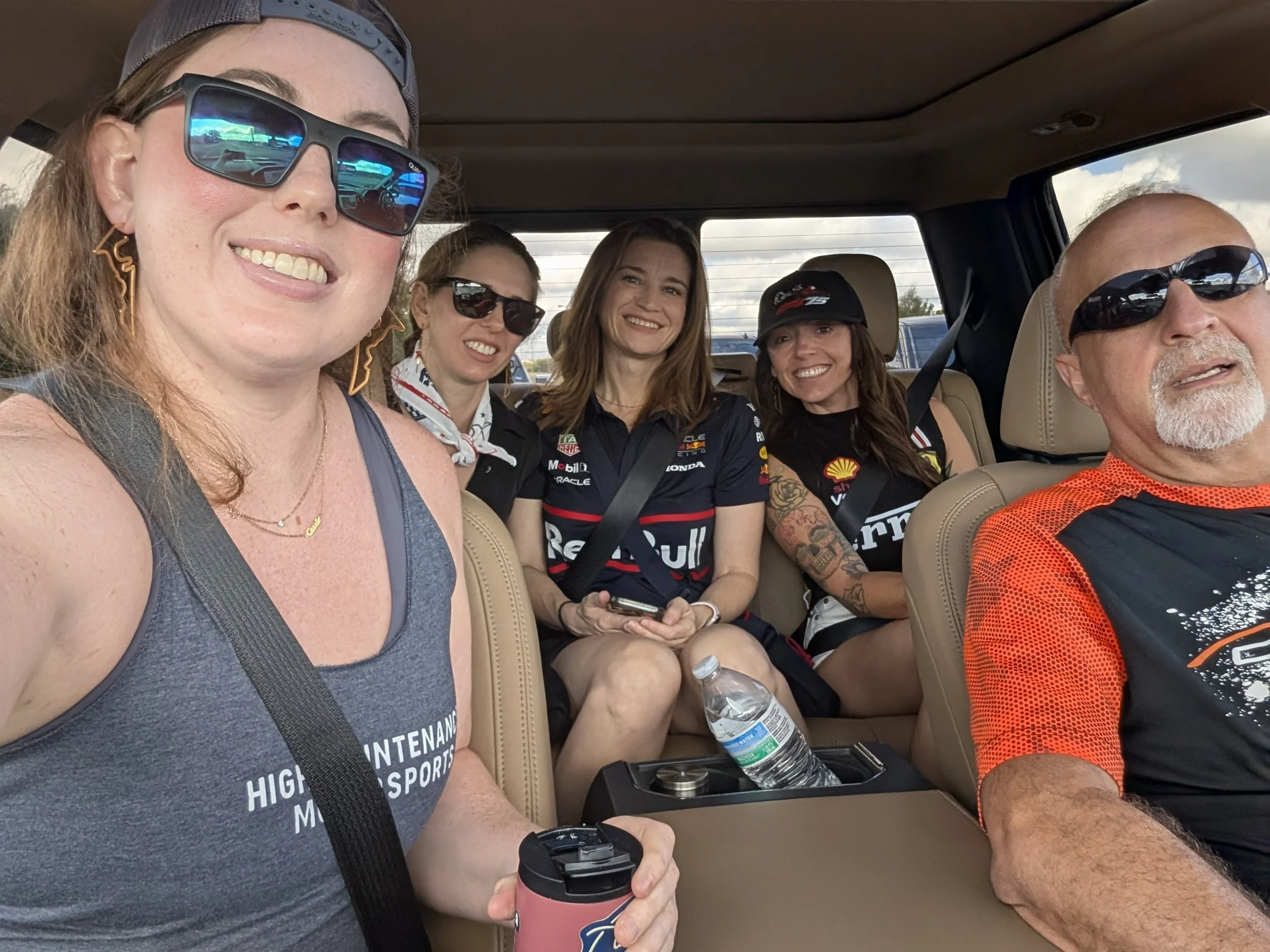 Group of five people smiling and sitting in a car, with three women and one man wearing sunglasses, and one woman holding a water bottle.