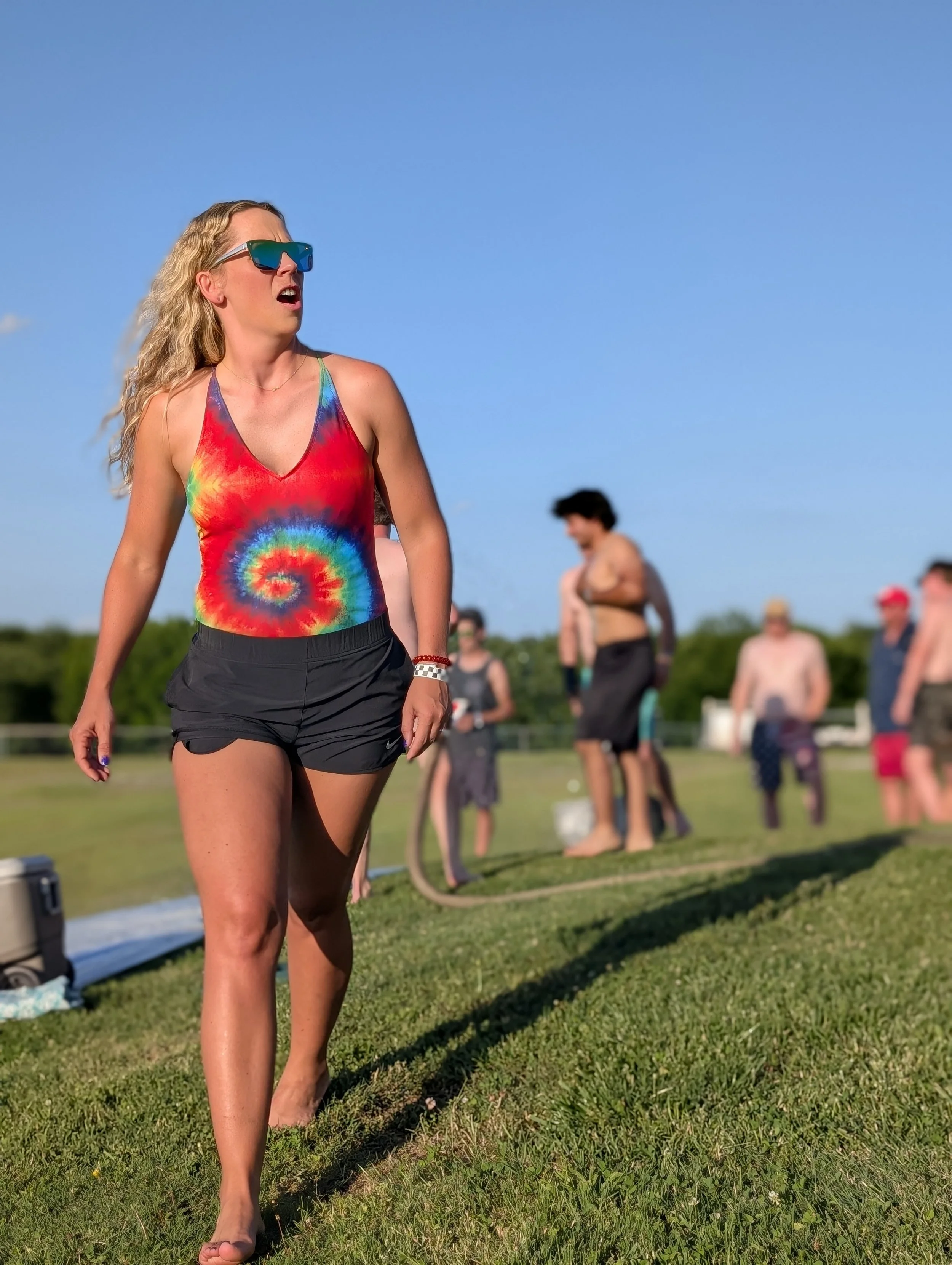 A woman with blonde wavy hair, wearing a colorful tie-dye tank top and black shorts, stands outdoors on grass with a group of people in the background, some shirtless, under a clear blue sky.