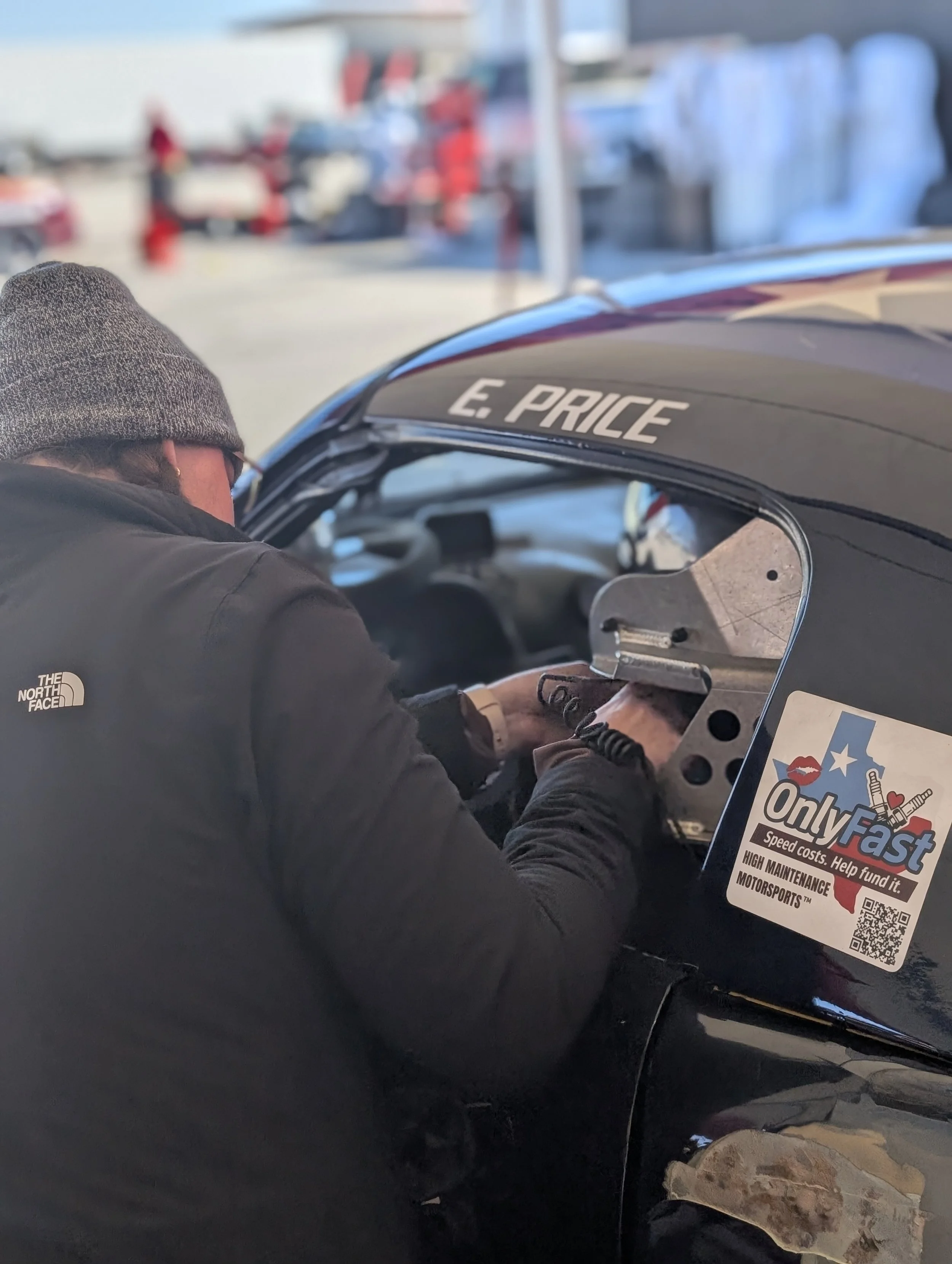A person wearing a gray beanie and black North Face jacket working on a race car with the name 'E. PRICE' on its windshield. The car has a sticker promoting high-maintenance motorsports and helping fund speed costs.