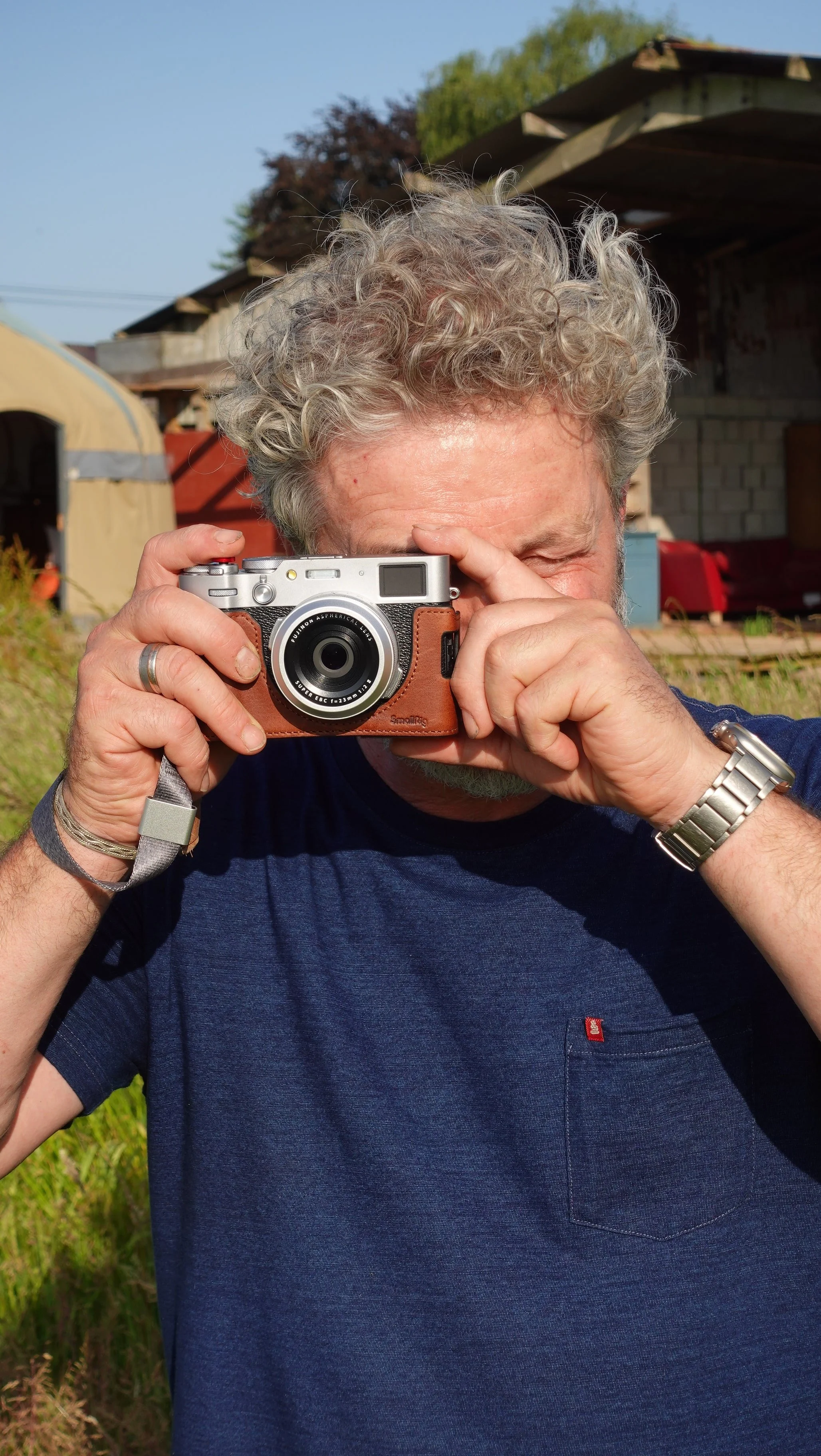 Older man with curly gray hair taking a photo with a camera outdoors on a sunny day.